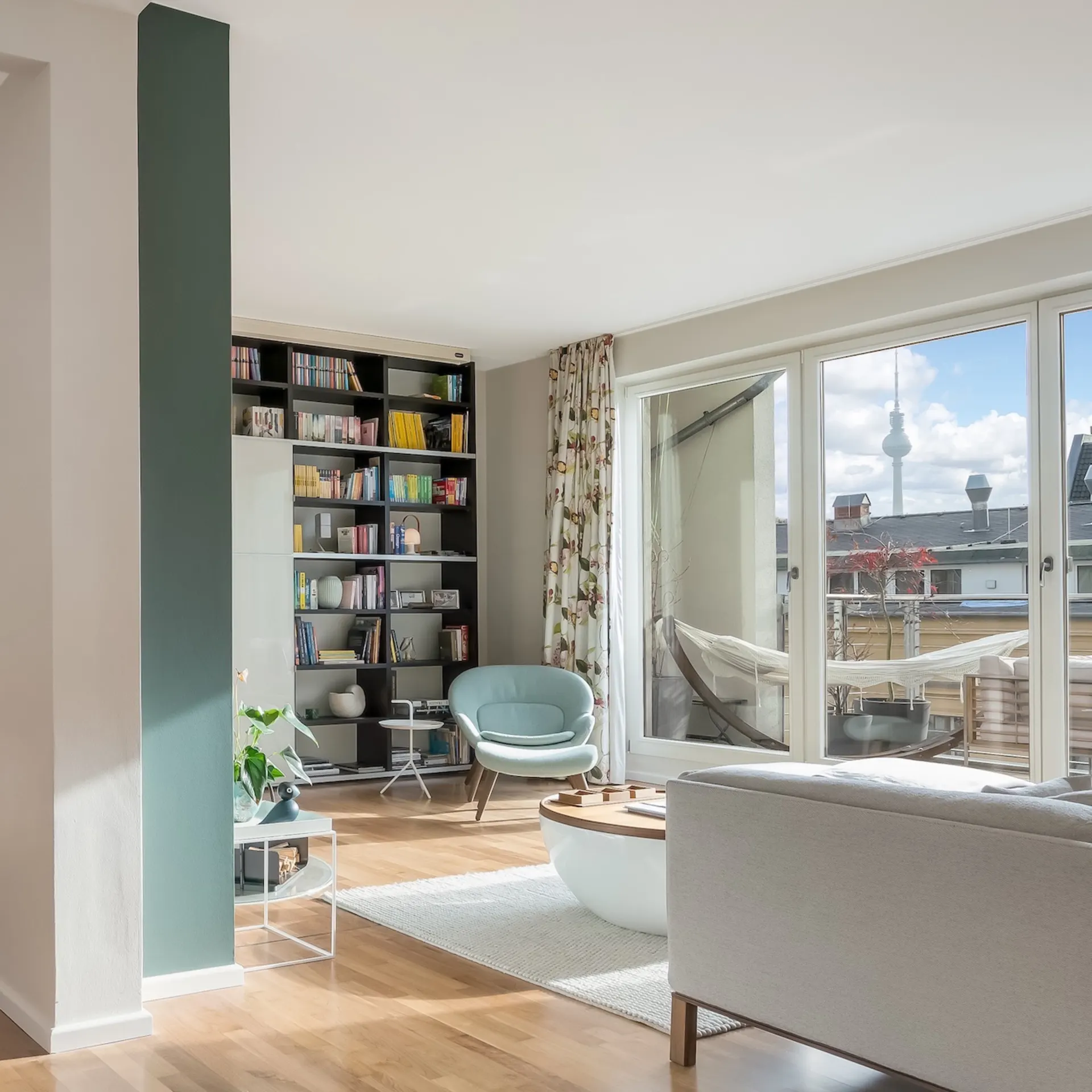 Helles Wohnzimmer mit Holzboden, Sessel, Regal, Sofa und großem Fenster zur Terrasse.