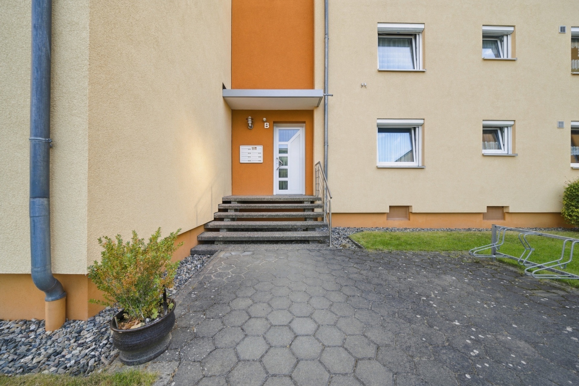 Eingang eines Mehrfamilienhauses mit Treppe, Briefkasten und Pflanzenkübel.