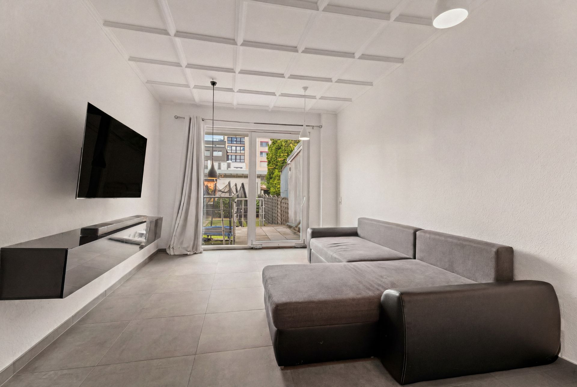 Modernes Wohnzimmer mit grauem Ecksofa, Wandfernseher und großen Fenstern zum Garten.
