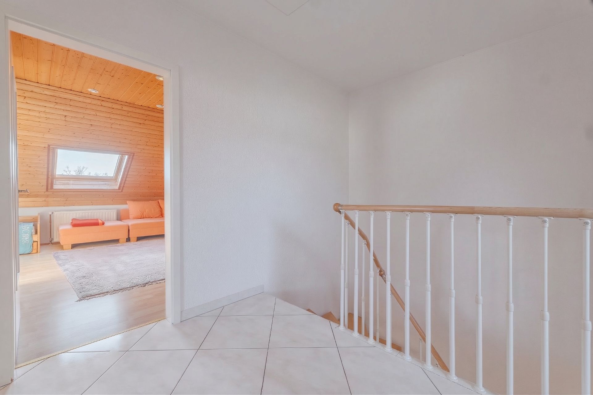 Helle Diele mit Treppengeländer und Blick in ein Zimmer mit Holzvertäfelung und Sofa.