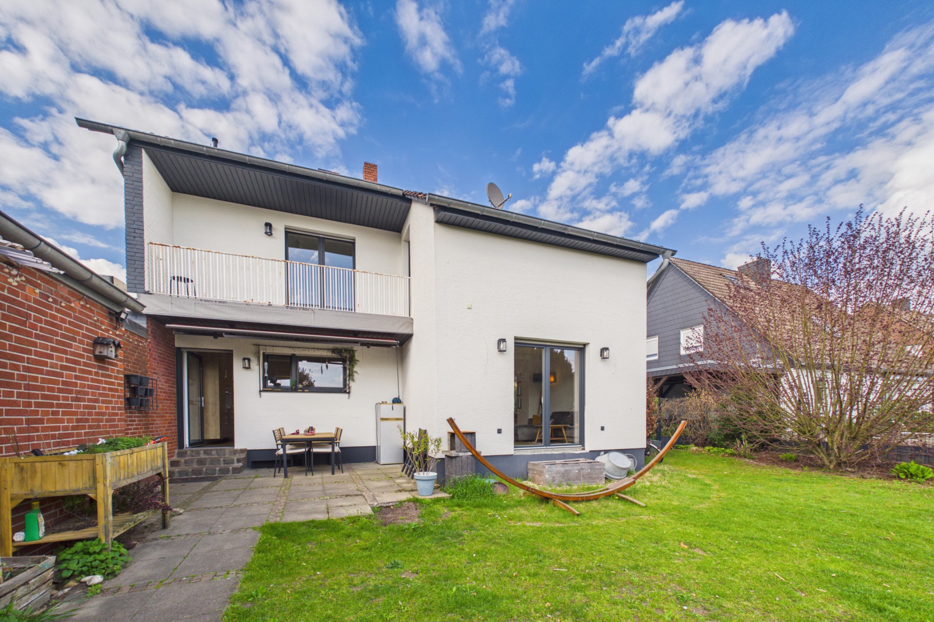 Zweistöckiges Haus mit Terrasse, Garten, Hängematte und Balkon bei Tageslicht.
