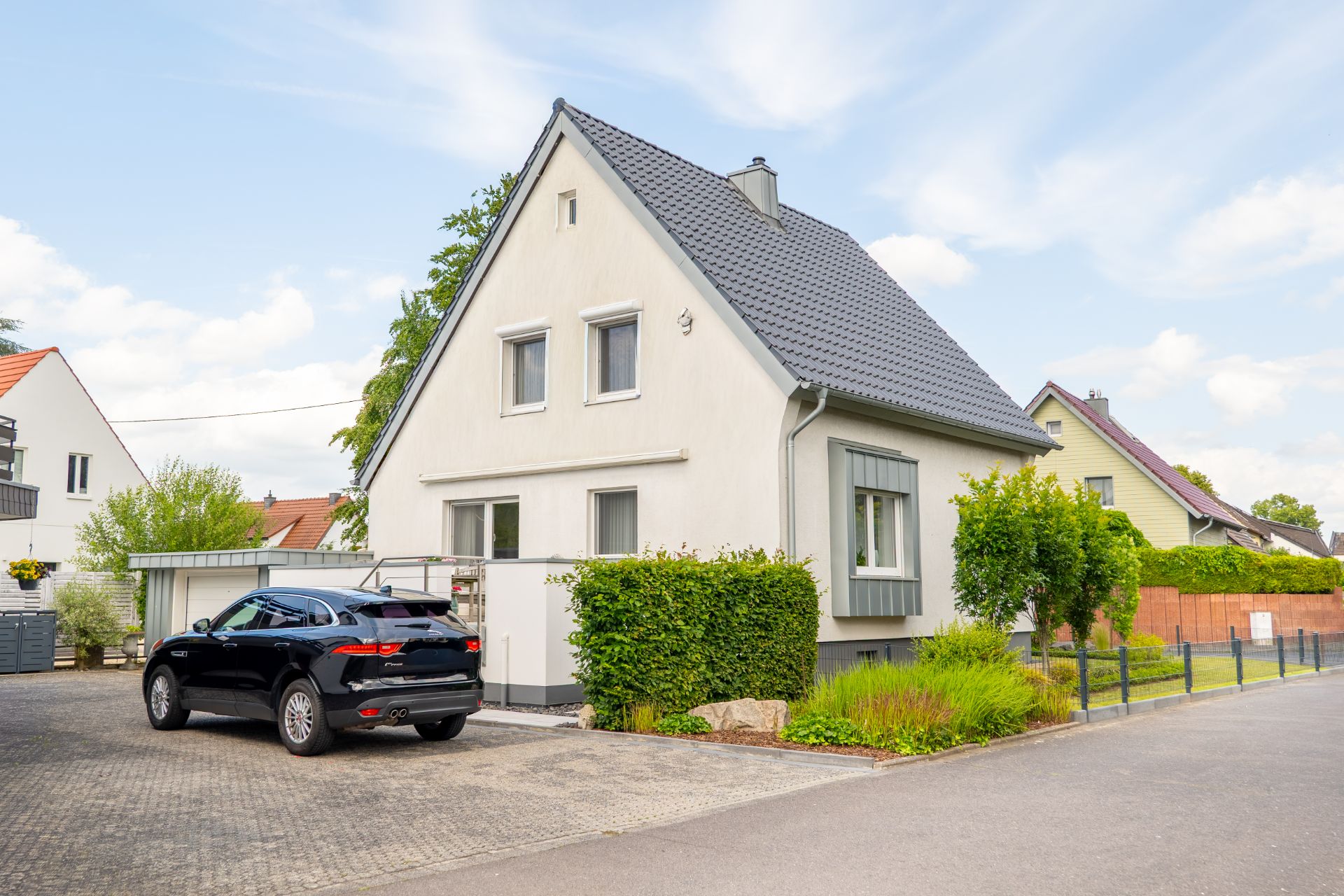 Modernes Einfamilienhaus mit grauem Dach und schwarzem Auto in Einfahrt, umgeben von Garten.