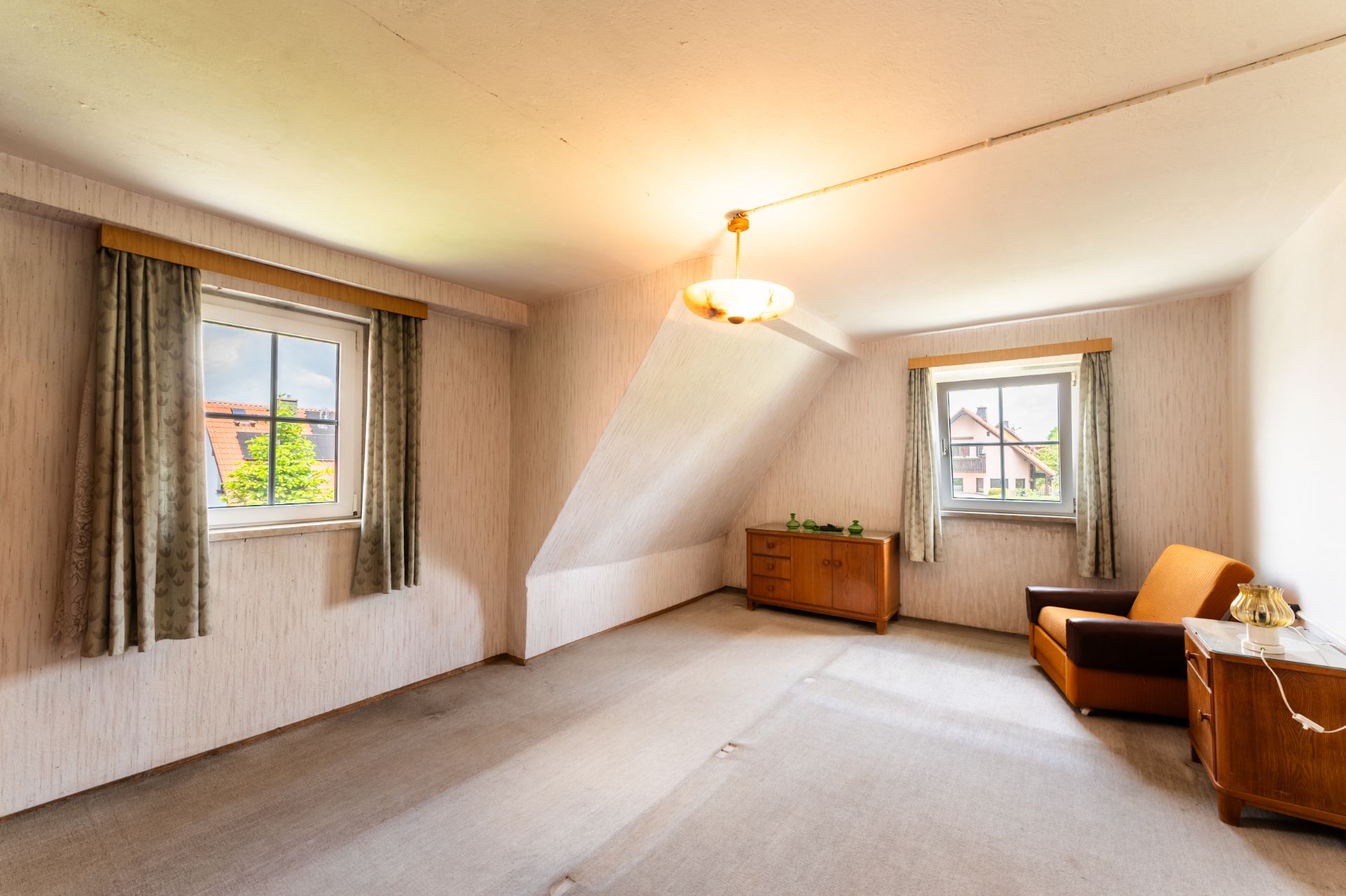 Helles Zimmer mit zwei Fenstern, Vorhängen, Sofa, zwei Kommoden und Deckenleuchte.
