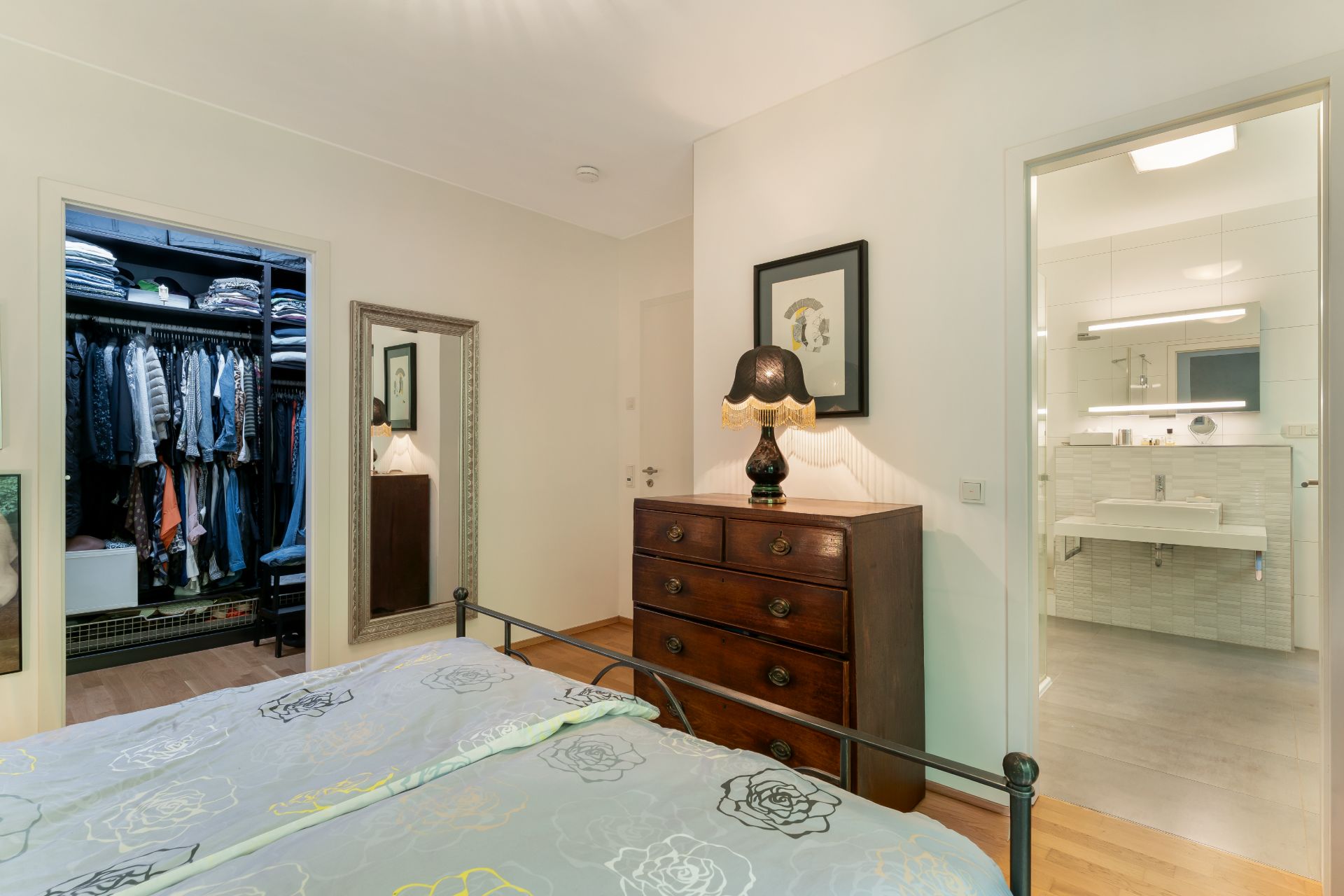 Schlafzimmer mit Bett, Kommode, Spiegel, offenem Kleiderschrank und Zugang zum Badezimmer.