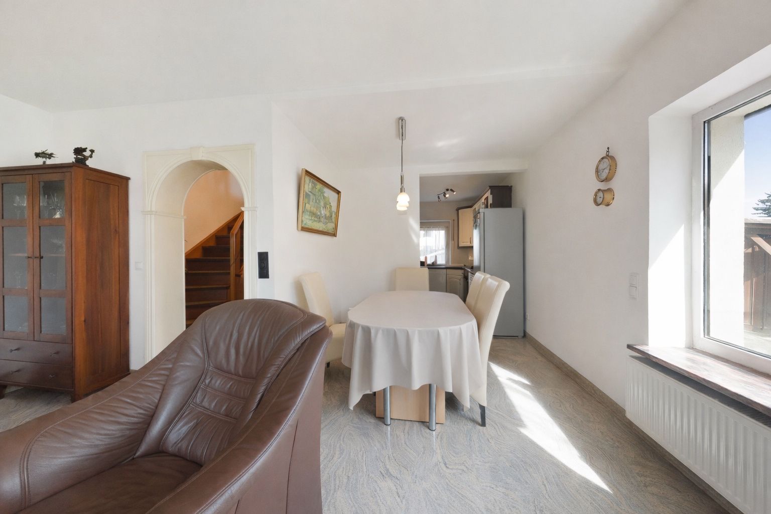 Helles Wohnzimmer mit braunem Sessel, Esstisch, Stühlen und Blick in die Küche.