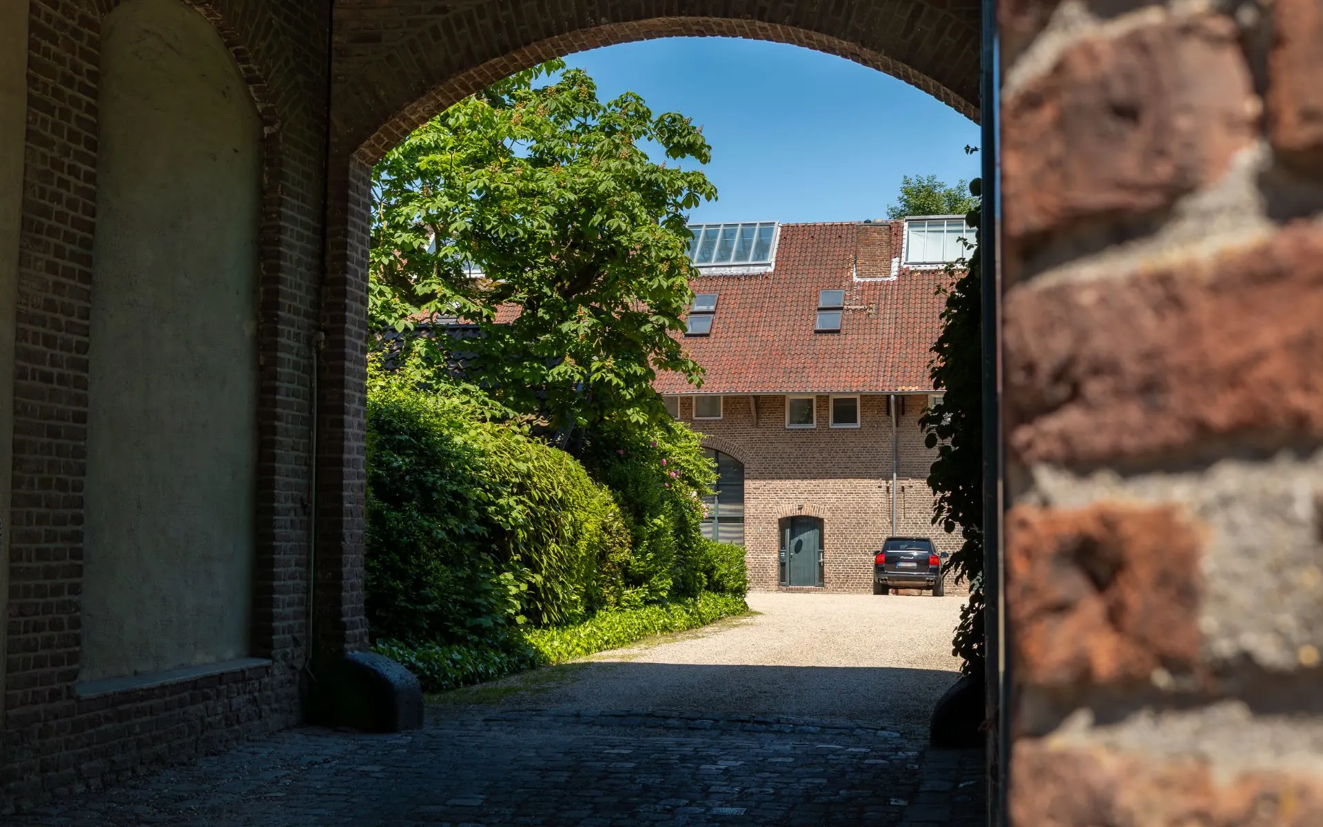 Blick durch einen Torbogen auf ein Wohnhaus mit rotem Dach und grünem Garten.