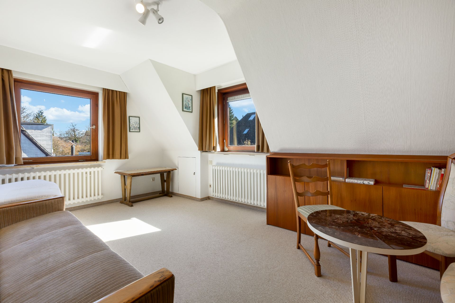 Helles Zimmer mit zwei Fenstern, einem Sofa, Tisch, Stuhl und Regal mit Büchern.