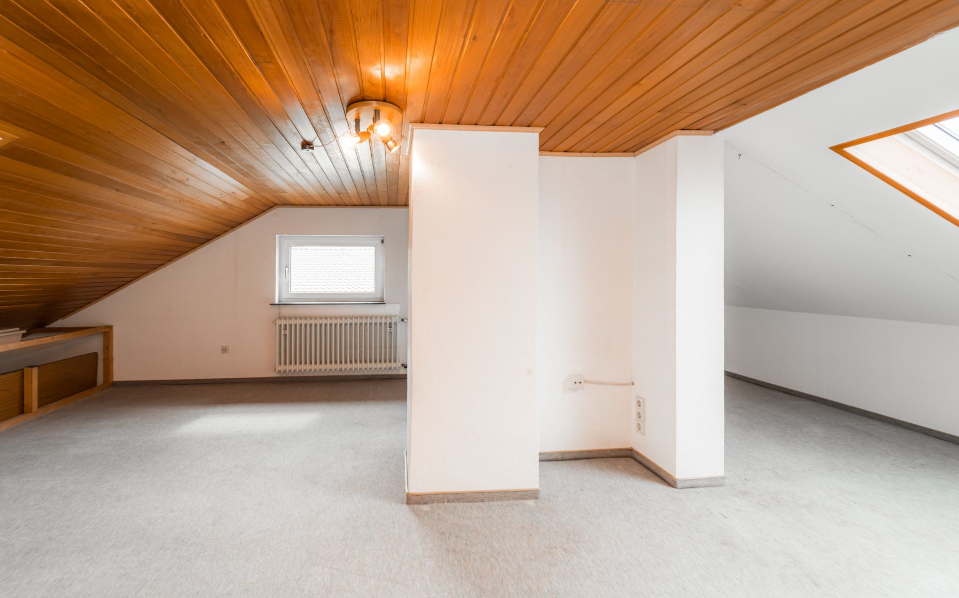 Helles Dachzimmer mit Holzdecke, Teppichboden, zwei weißen Wänden und Dachfenster.