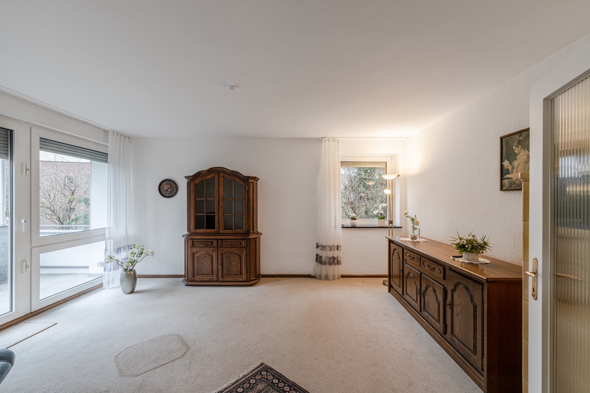 Helles Wohnzimmer mit großen Fenstern, Vitrine und Sideboard aus Holz, Teppich und Dekoration.