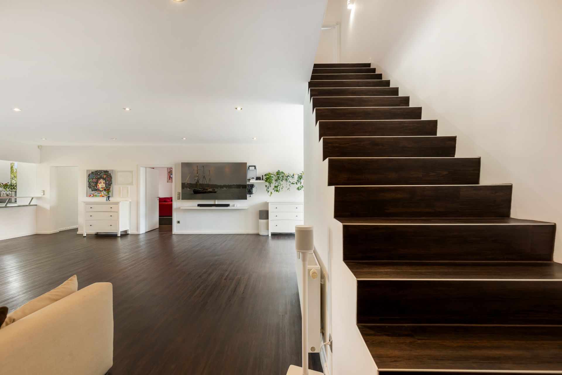 Modernes Wohnzimmer mit dunklem Holzboden und Treppe, TV an der Wand, weiße Wände und Möbel.