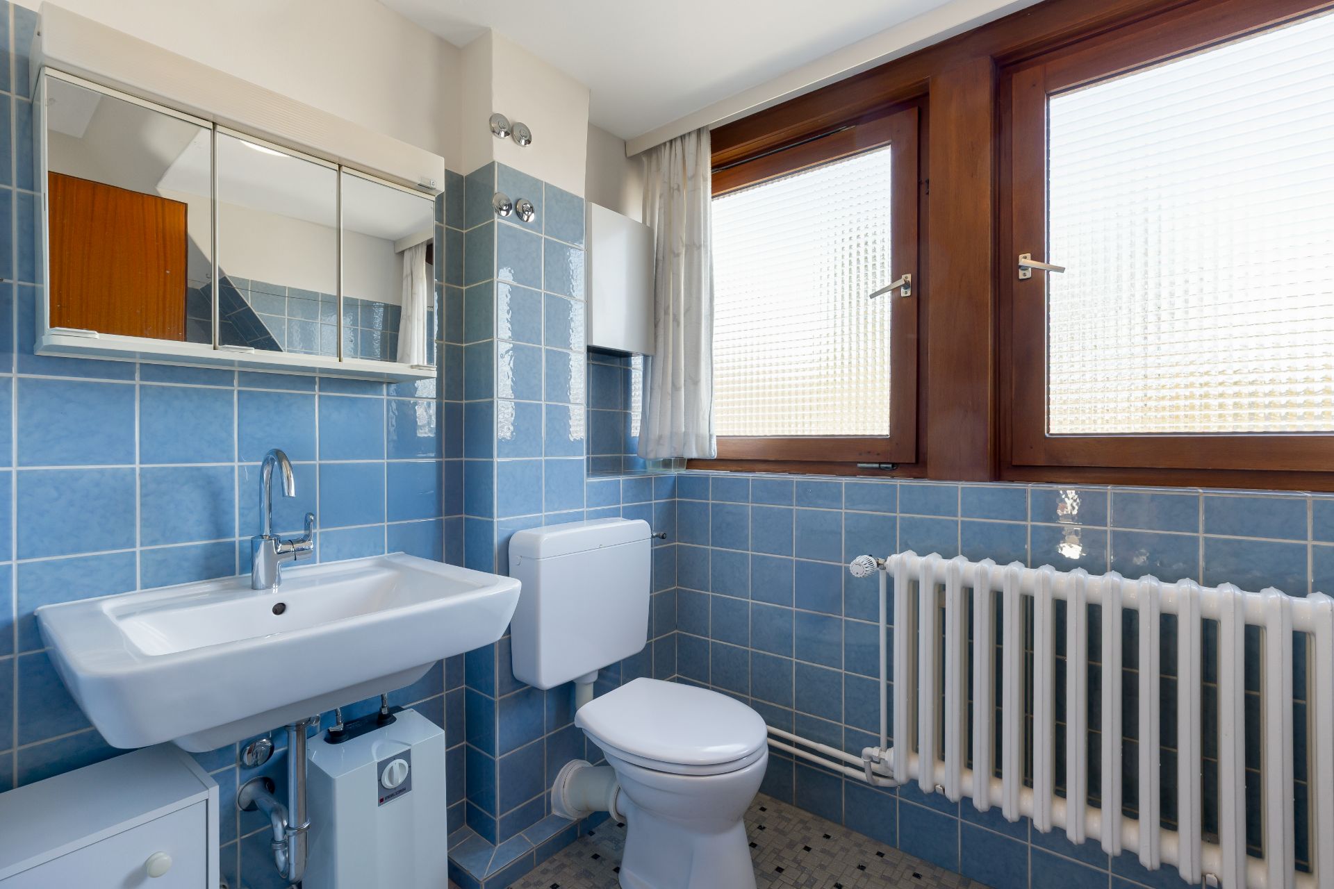 Kleines Badezimmer mit blauen Fliesen, Waschbecken, WC, Heizkörper und Fenster.
