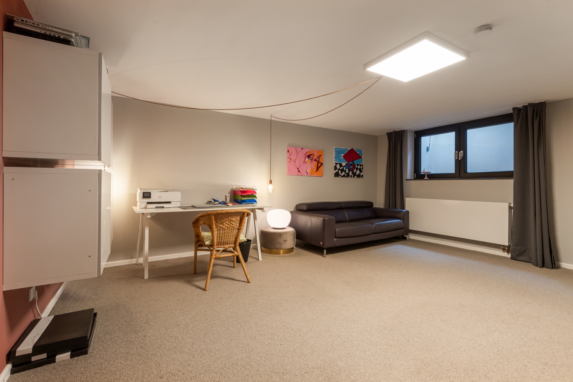 Helles Zimmer mit Sofa, Schreibtisch, Stuhl, Wandregalen und zwei Fenstern mit Vorhängen.