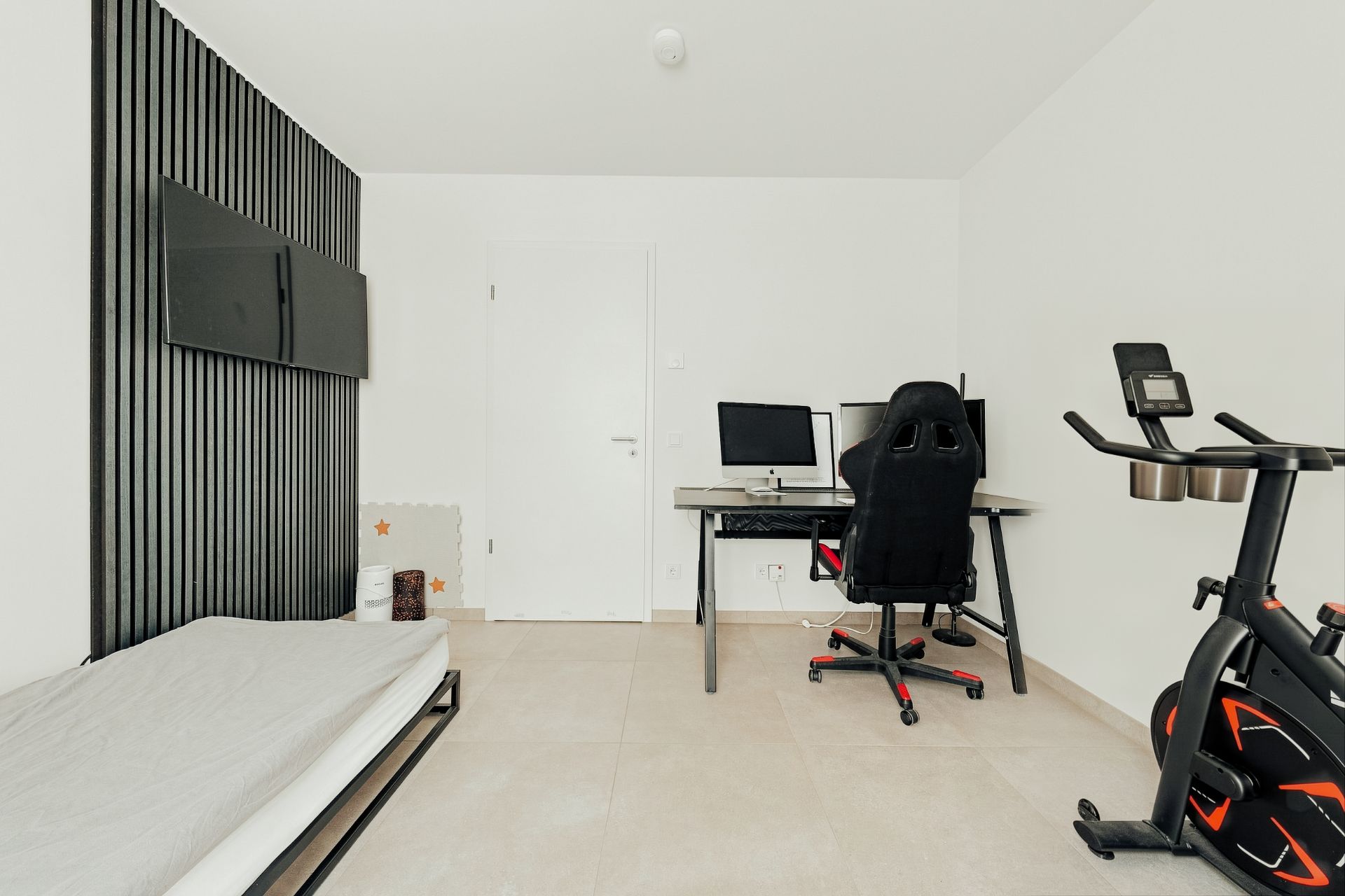 Helles Zimmer mit Bett, Schreibtisch, Computer, Drehstuhl und Heimtrainer auf Fliesenboden.