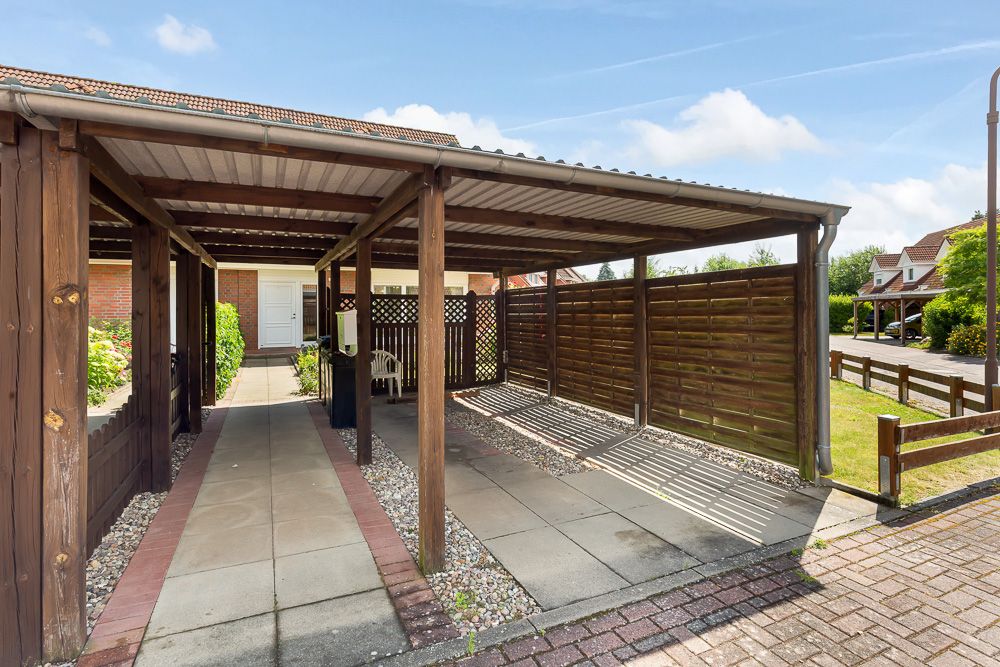Überdachter Carport mit Holzwänden und gepflastertem Boden vor einem Wohnhaus.