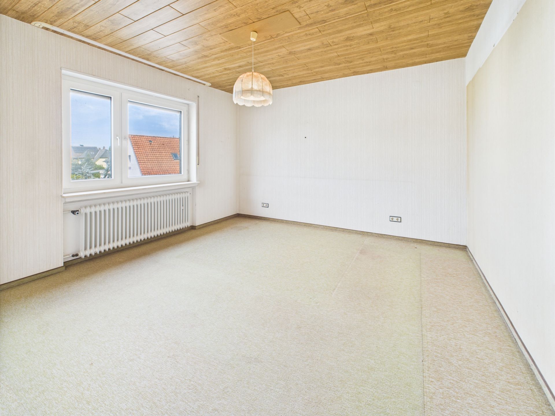 Leeres Zimmer mit Teppichboden, großem Fenster, Holzdecke und Hängelampe.