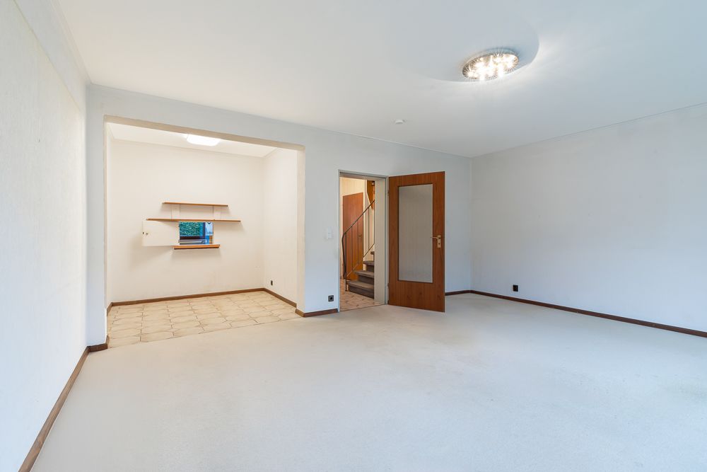 Helles, leeres Wohnzimmer mit Tür und Zugang zu einem gefliesten Bereich mit Wandregal und kleinem Fenster.