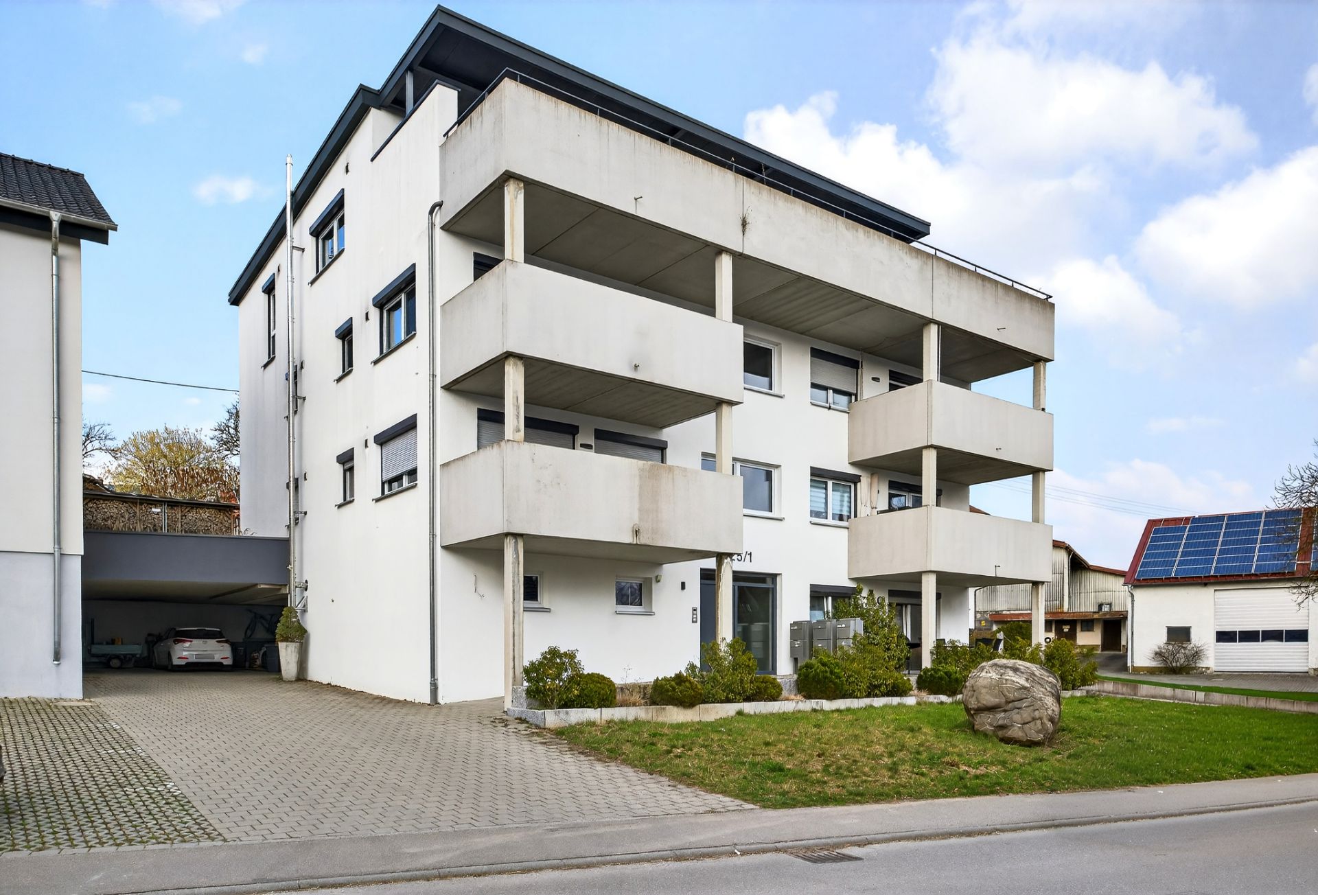 Modernes, weißes Mehrfamilienhaus mit Balkonen und Carport, davor eine kleine Grünfläche.