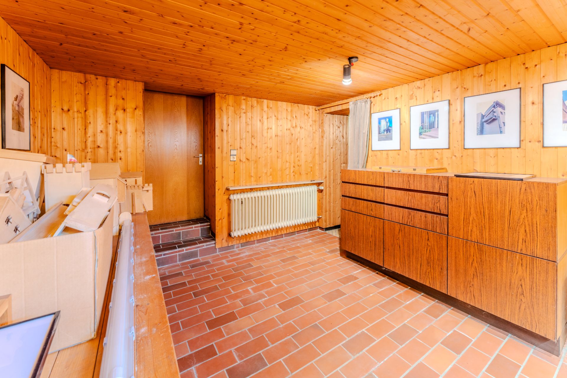 Holzverkleideter Raum mit Kommode, Heizkörper, Fliesenboden und gerahmten Bildern an der Wand.