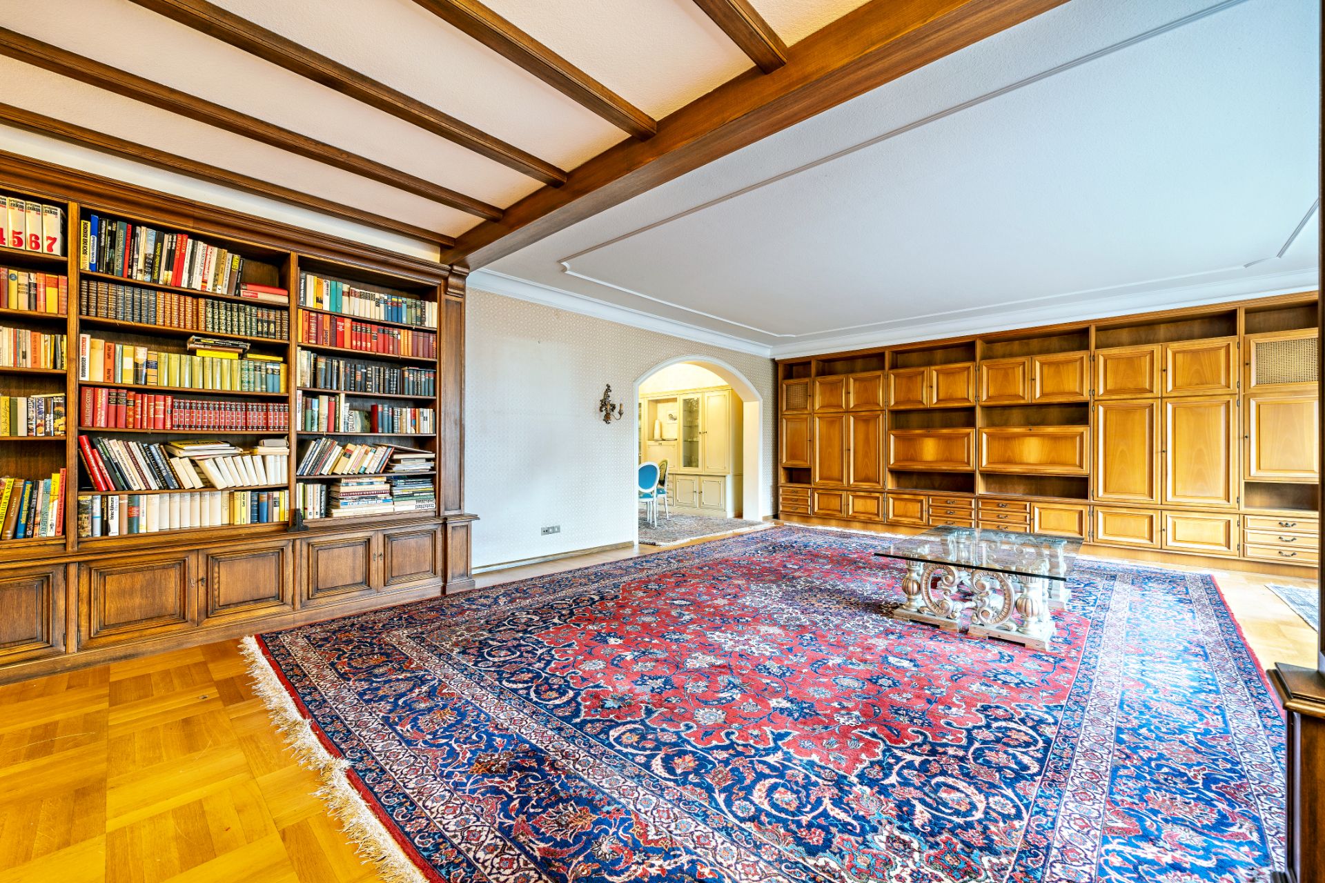 Helles Wohnzimmer mit großem Teppich, Bücherregal, Holzschränken und Glastisch.