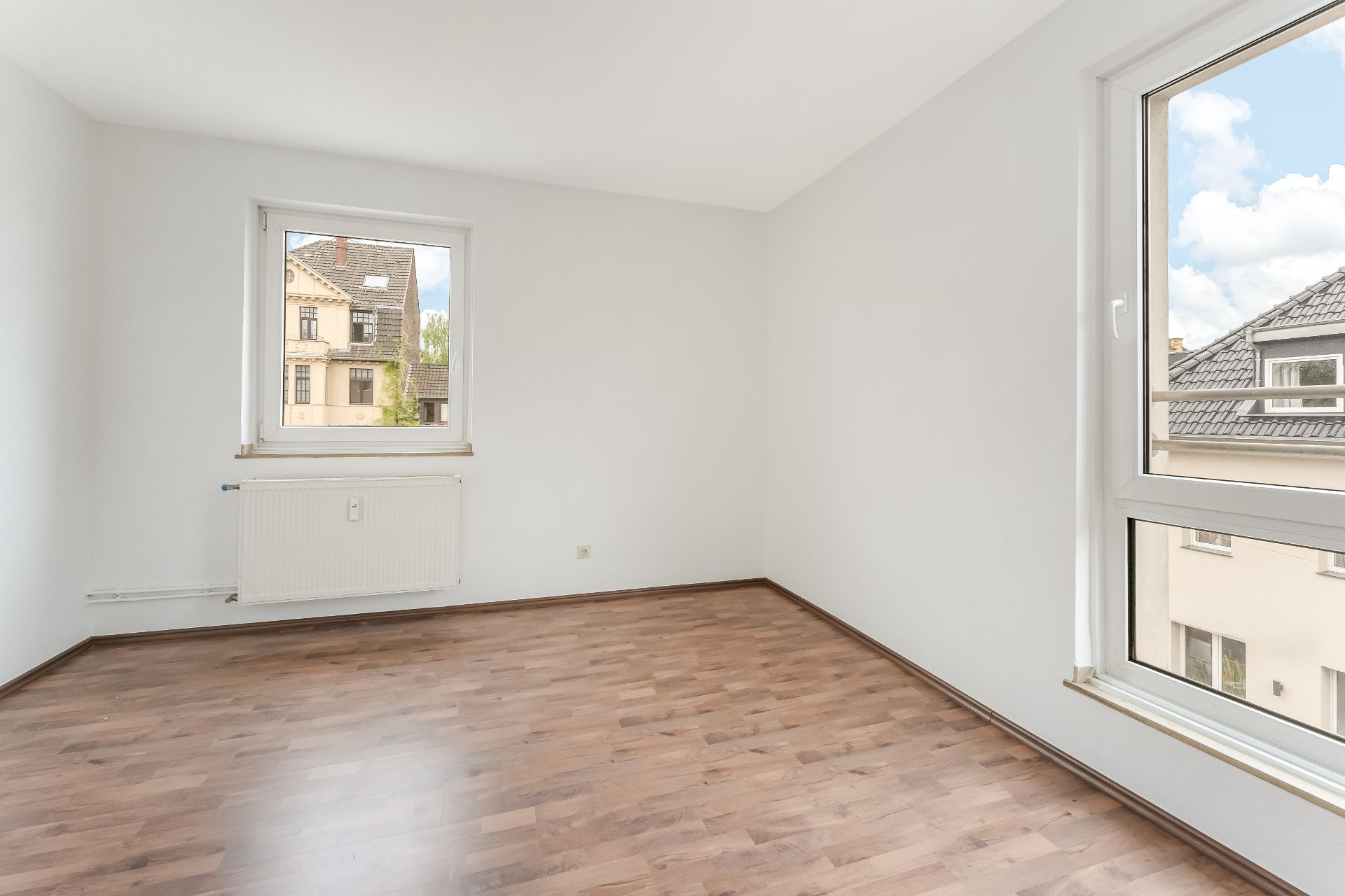Heller, leerer Raum mit Holzfußboden, weißen Wänden und zwei Fenstern.