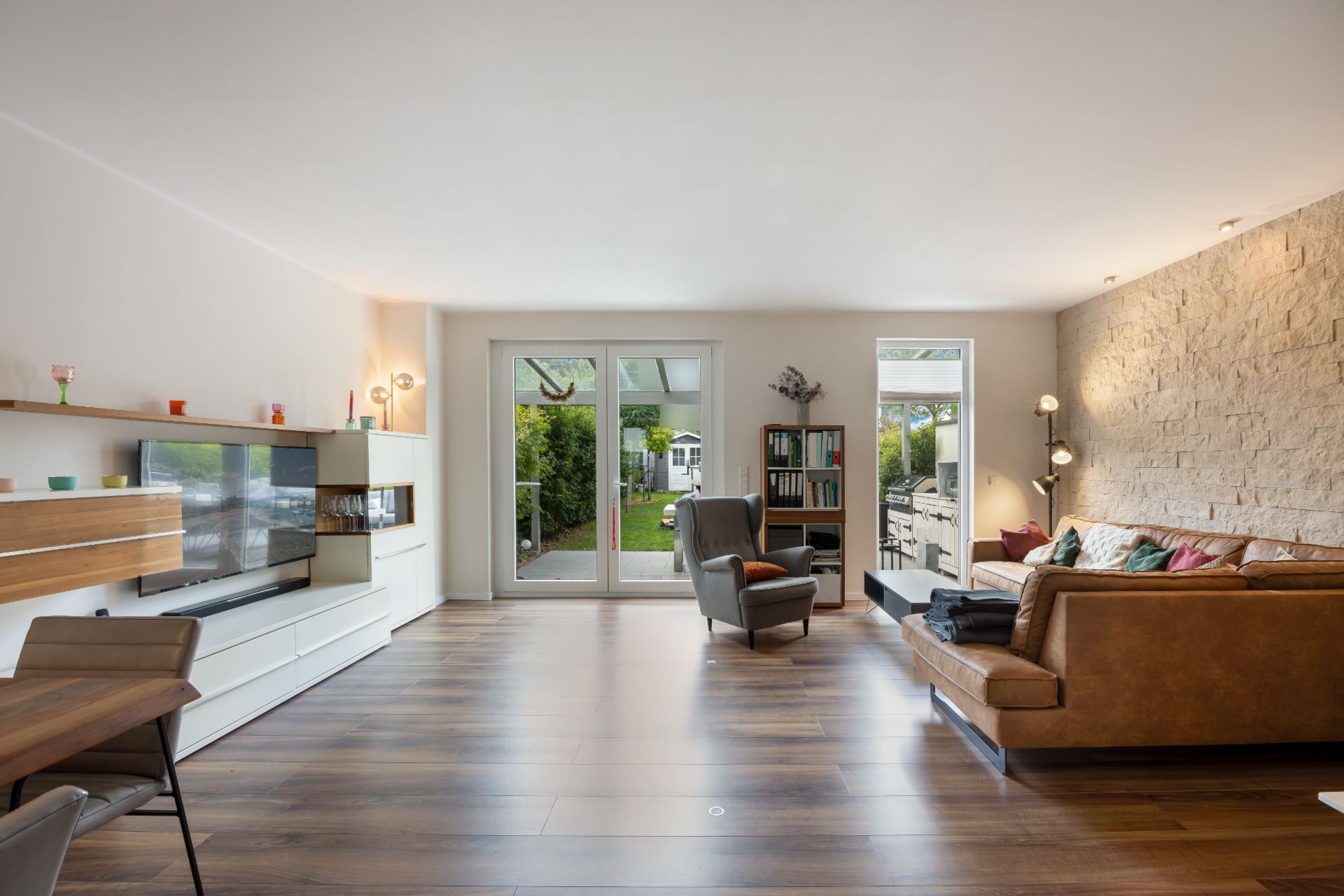 Helles Wohnzimmer mit braunem Sofa, Fernseher, Sesseln und Zugang zum Garten.