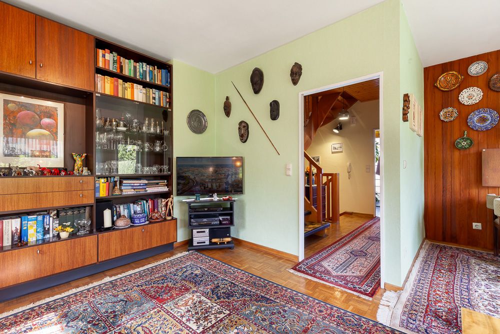 Wohnzimmer mit Bücherregal, Fernseher, Teppichen und Wanddekorationen, Blick auf Flur mit Treppe.