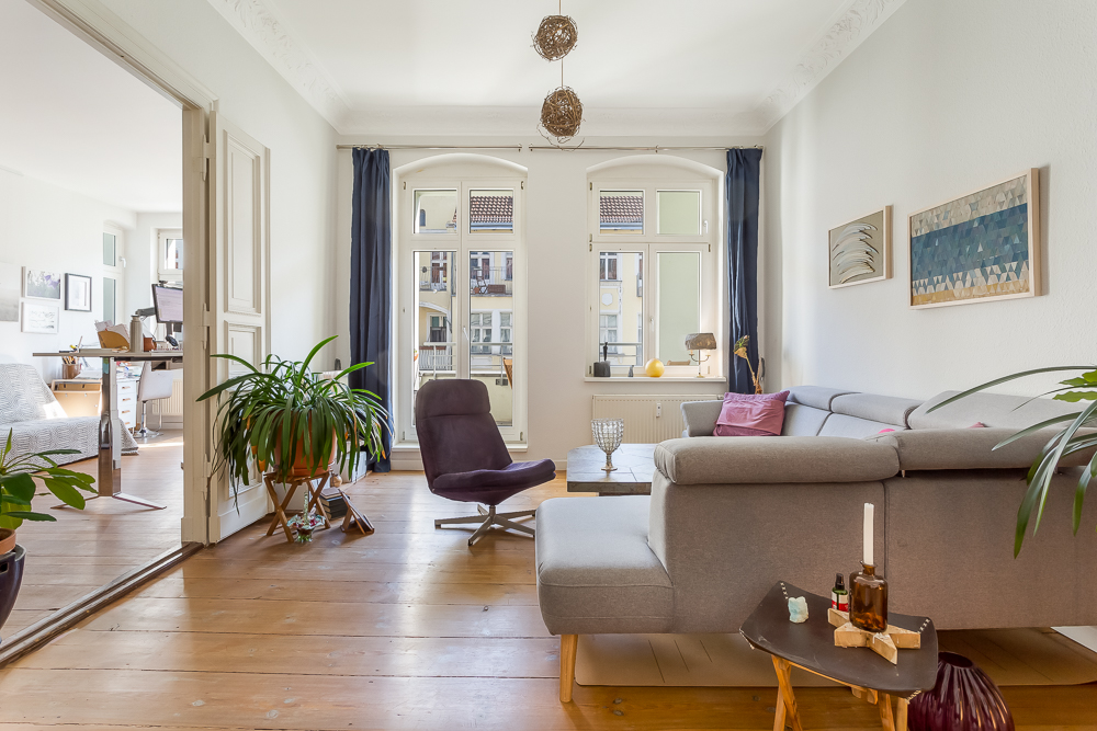 Helles Wohnzimmer mit grauem Sofa, Sessel, Pflanzen und Holzboden, Zugang zu Arbeitszimmer.