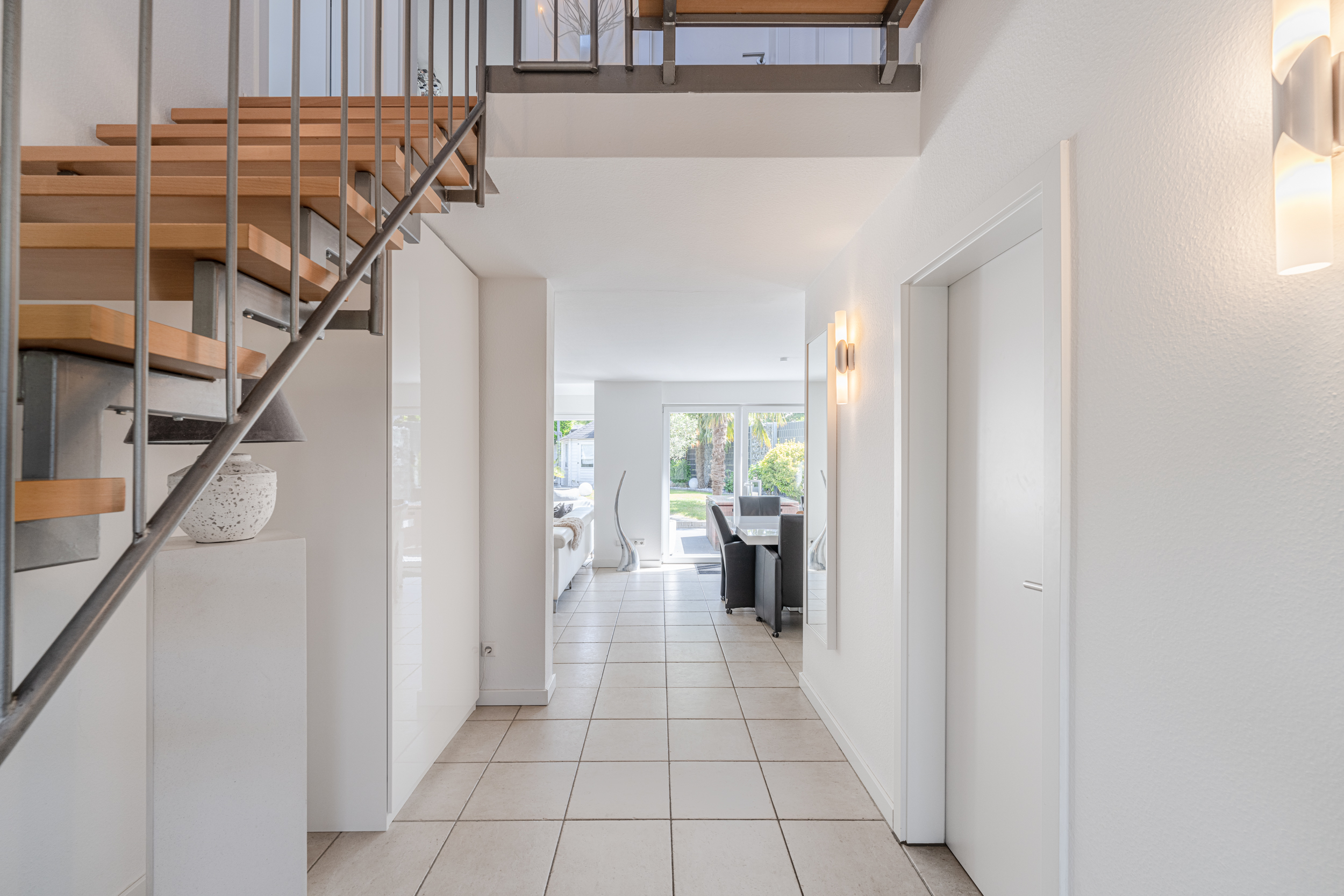 Heller Flur mit Treppe, Fliesenboden und Blick ins Wohnzimmer mit Glasfront zum Garten.