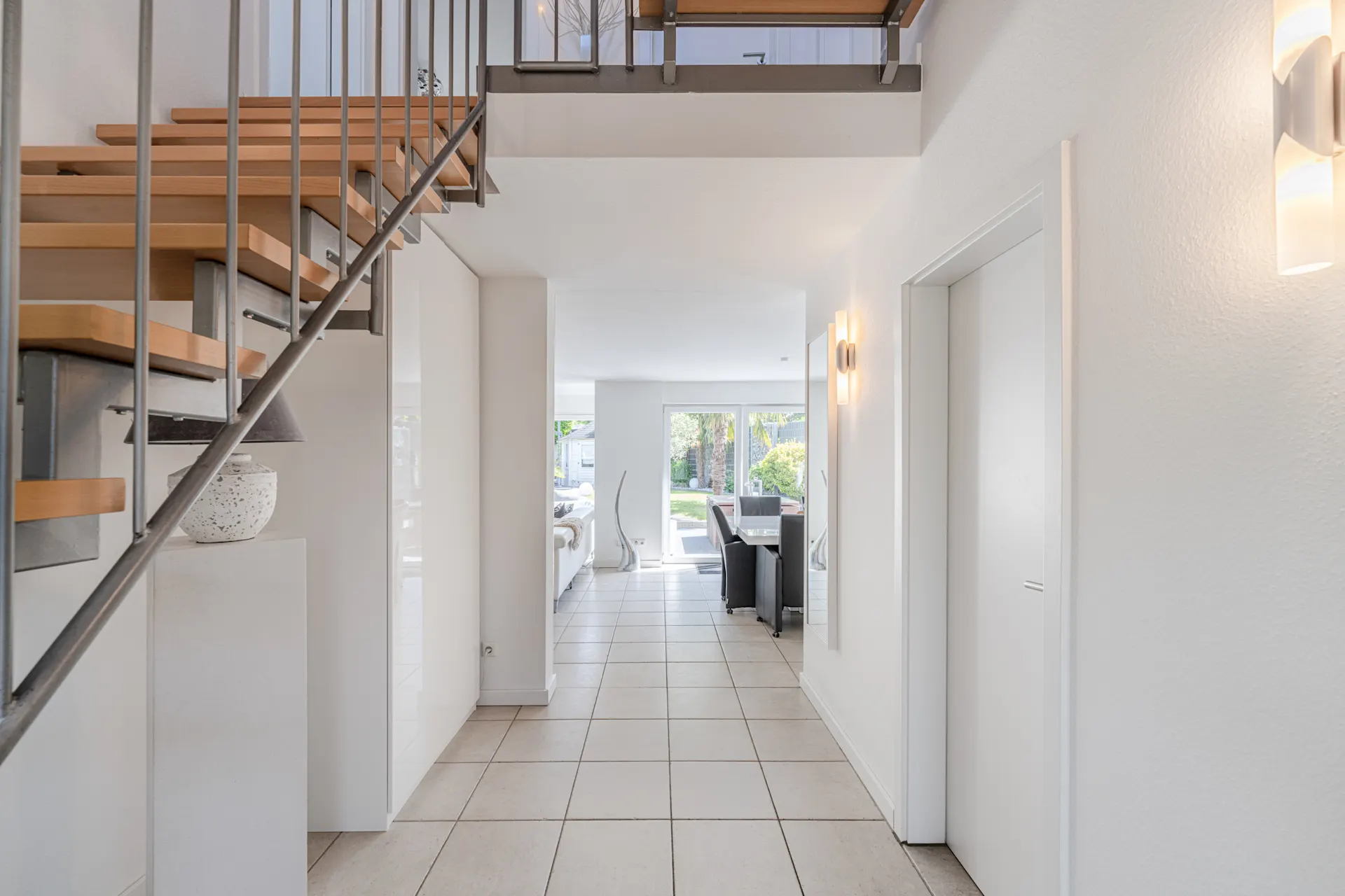 Heller Flur mit Treppe, Fliesenboden und Blick ins Wohnzimmer mit Glasfront zum Garten.