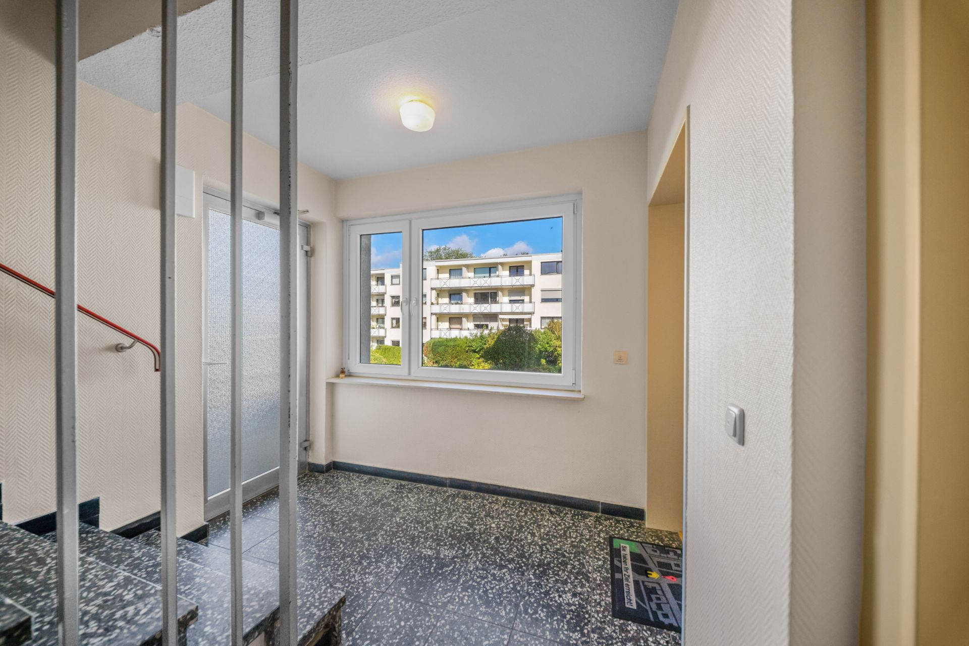 Heller Hausflur mit Fenster, Blick auf Wohngebäude und Treppengeländer.