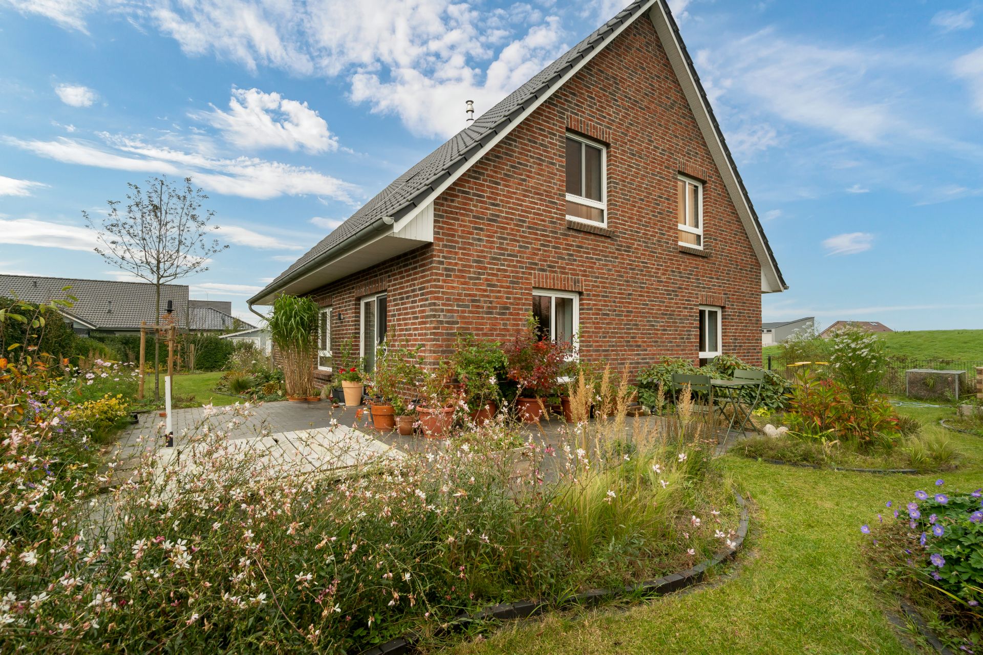 Backsteinhaus mit Garten, Terrasse und Blumenbeeten an einem sonnigen Tag.