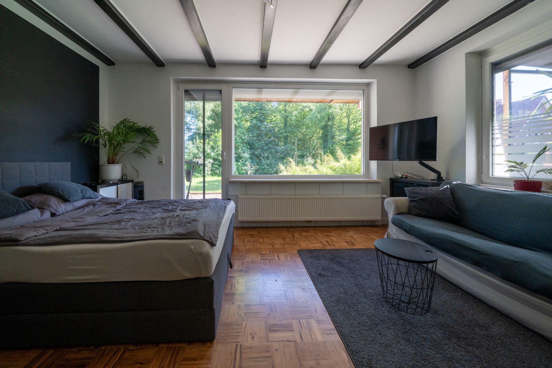 Helles Schlafzimmer mit Bett, Sofa, großem Fenster und Fernseher auf Holzfußboden.