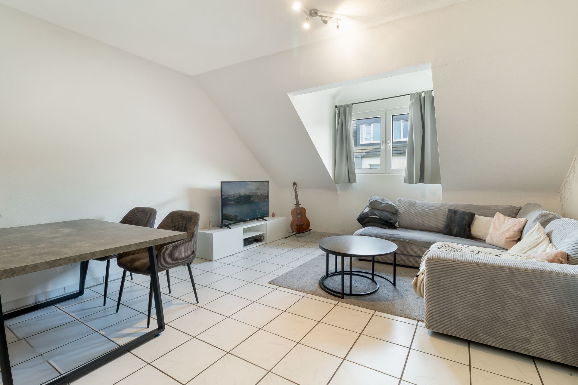 Helles Wohnzimmer mit Ecksofa, Couchtisch, Esstisch, Stühlen, Fernseher und Gitarre am Fenster.