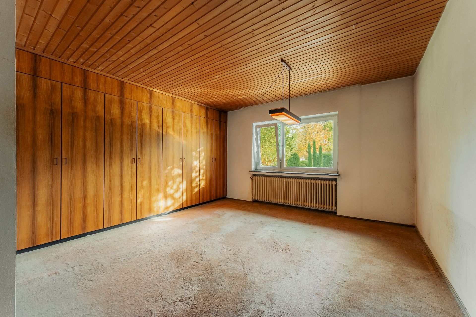 Leeres Zimmer mit Holzdecke, Einbauschränken und Fenster mit Blick nach draußen.