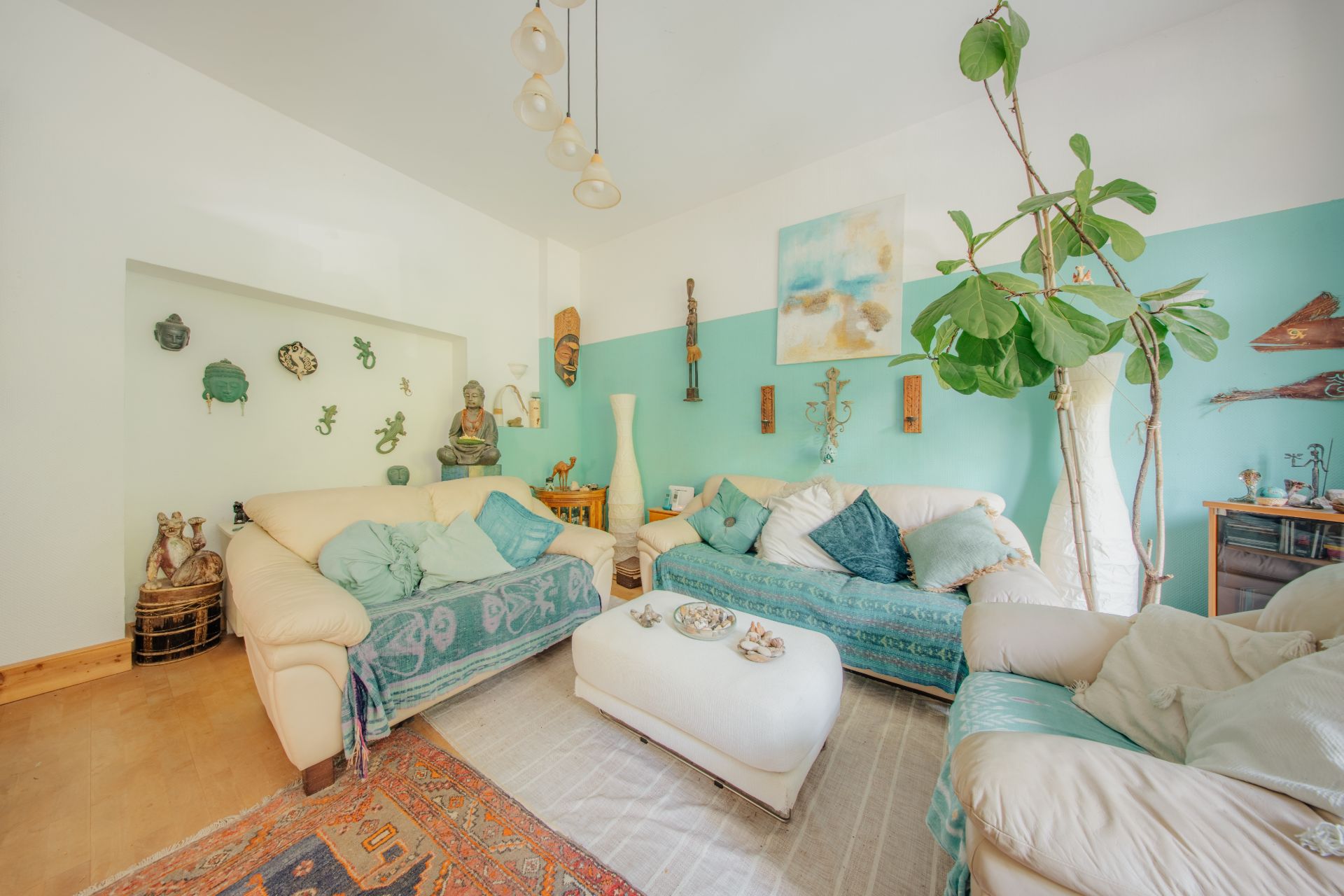Helles Wohnzimmer mit drei Sofas, großem grünen Baum, Wanddekorationen und Couchtisch.