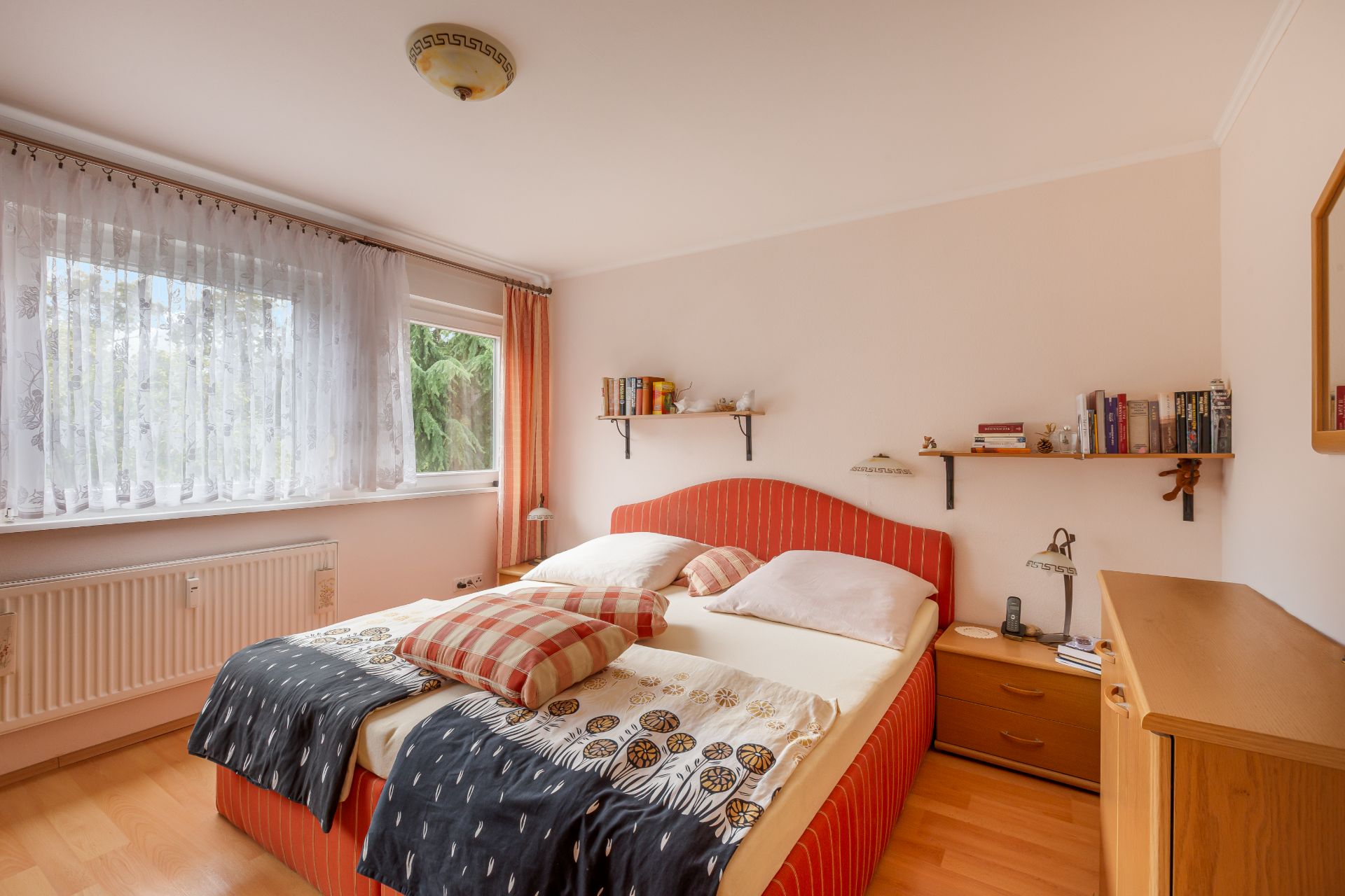 Schlafzimmer mit Doppelbett, Nachttisch, Bücherregalen und großem Fenster mit Gardine.