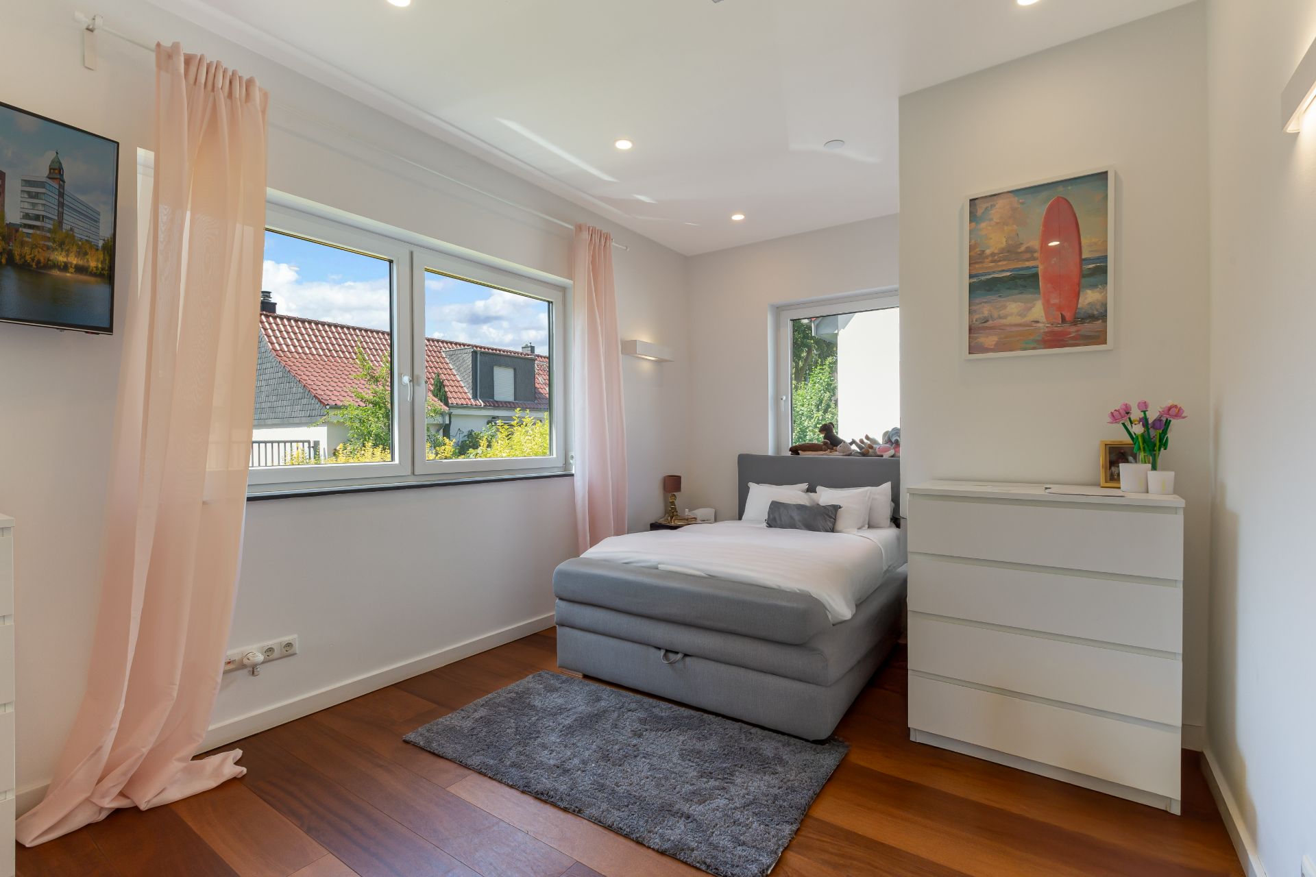 Helles Schlafzimmer mit großem Fenster, grauem Bett, Teppich und weißer Kommode.