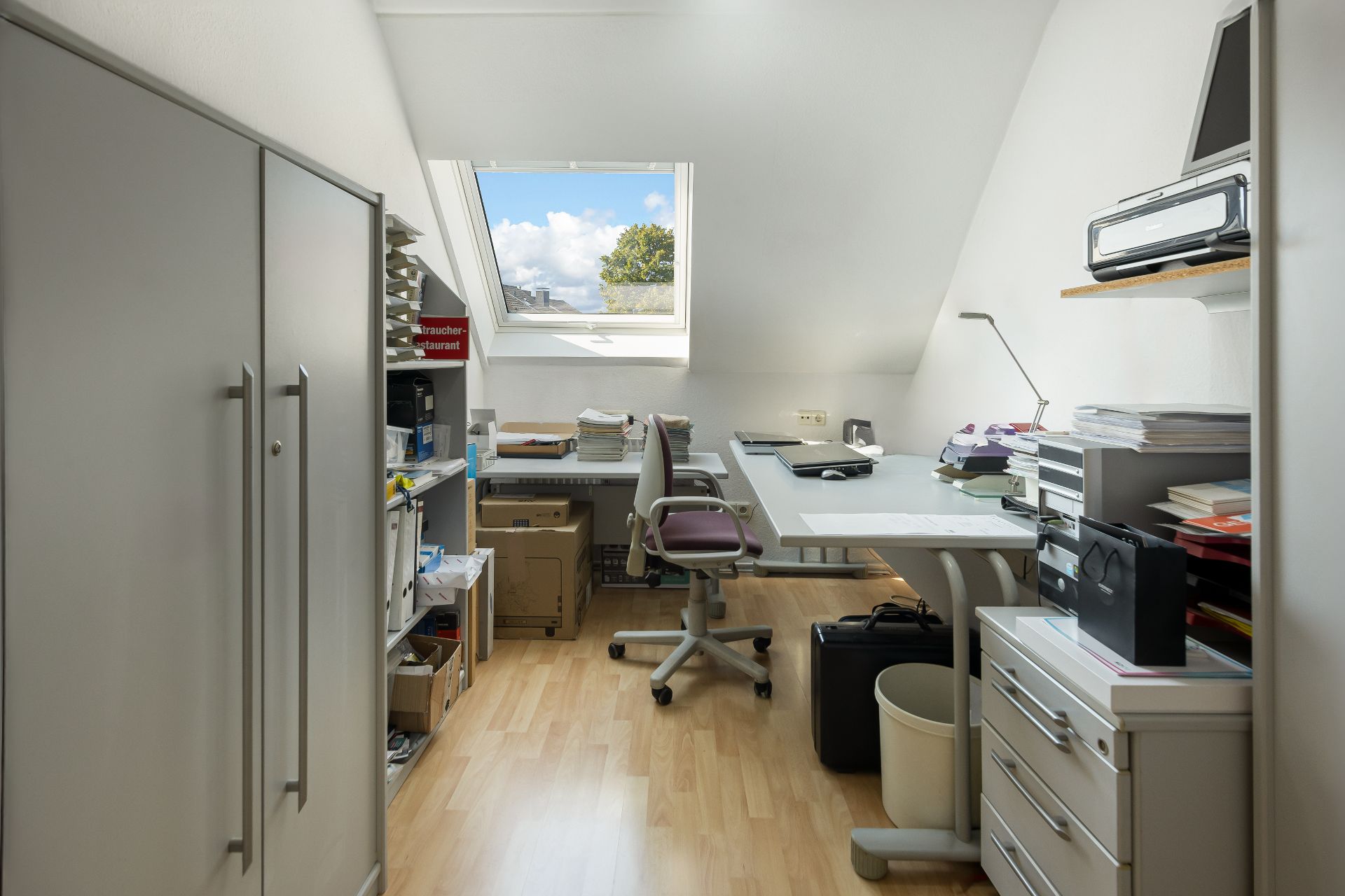 Helles Arbeitszimmer mit Schreibtisch, Stuhl, Regalen und Dachfenster.