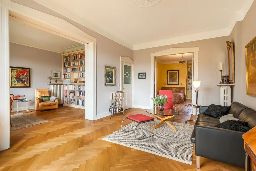 Helles Wohnzimmer mit Holzboden, schwarzem Sofa, Sessel, Bücherregal und Blick in ein Schlafzimmer.
