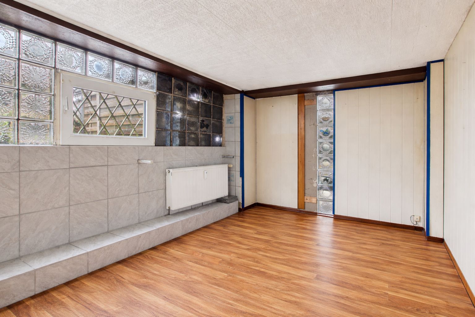 Leerer Raum mit Holzfußboden, Glasbausteinfenster und Heizung an der Wand.