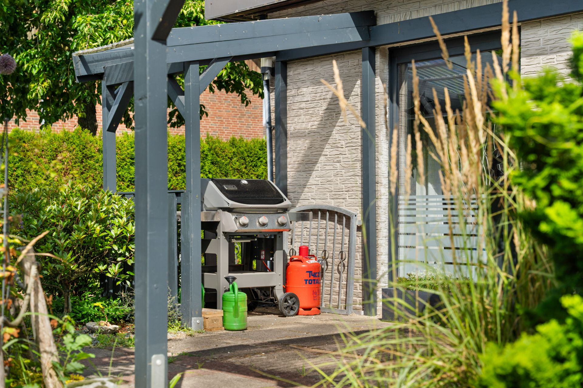 Gasgrill, Gasflaschen und Gießkanne stehen auf einer Terrasse neben einem Haus.