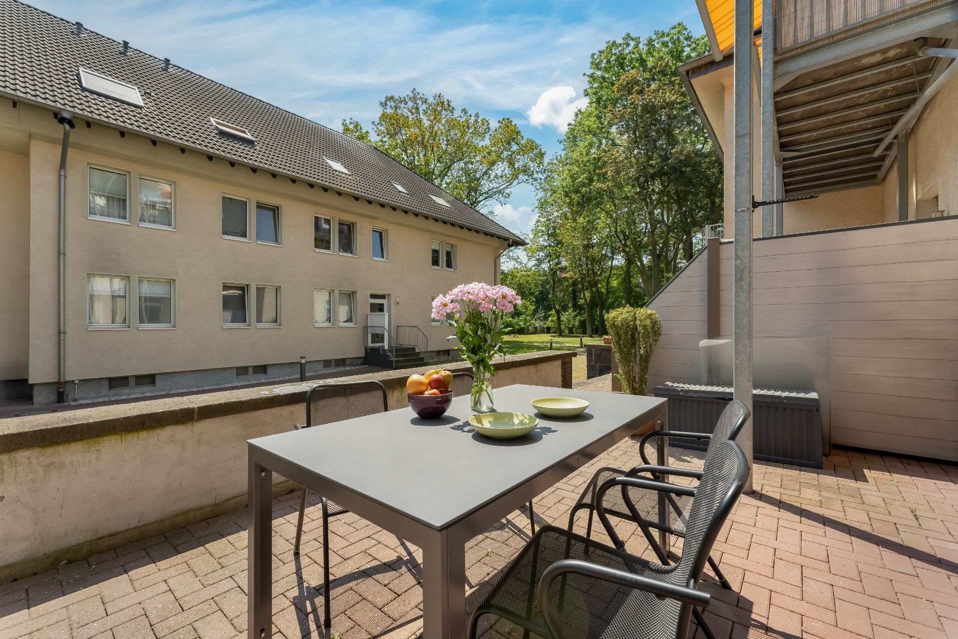 Terrasse mit Tisch, Stühlen, Blumen und Obstschale, Blick auf Wohnhaus und grüne Bäume.