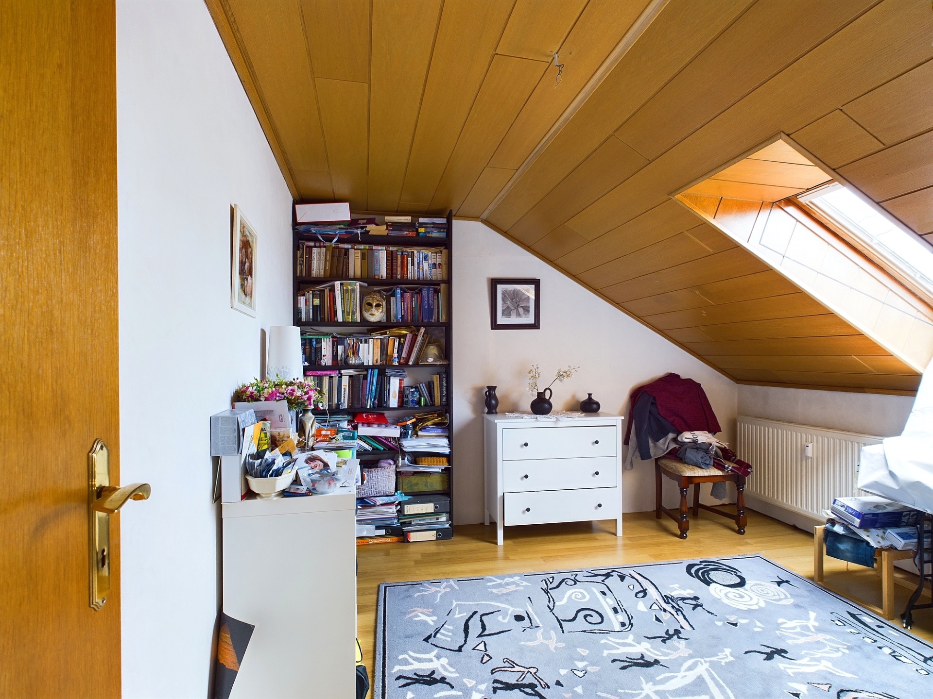 Dachzimmer mit Bücherregal, Kommode, Teppich und Schrägfenster.