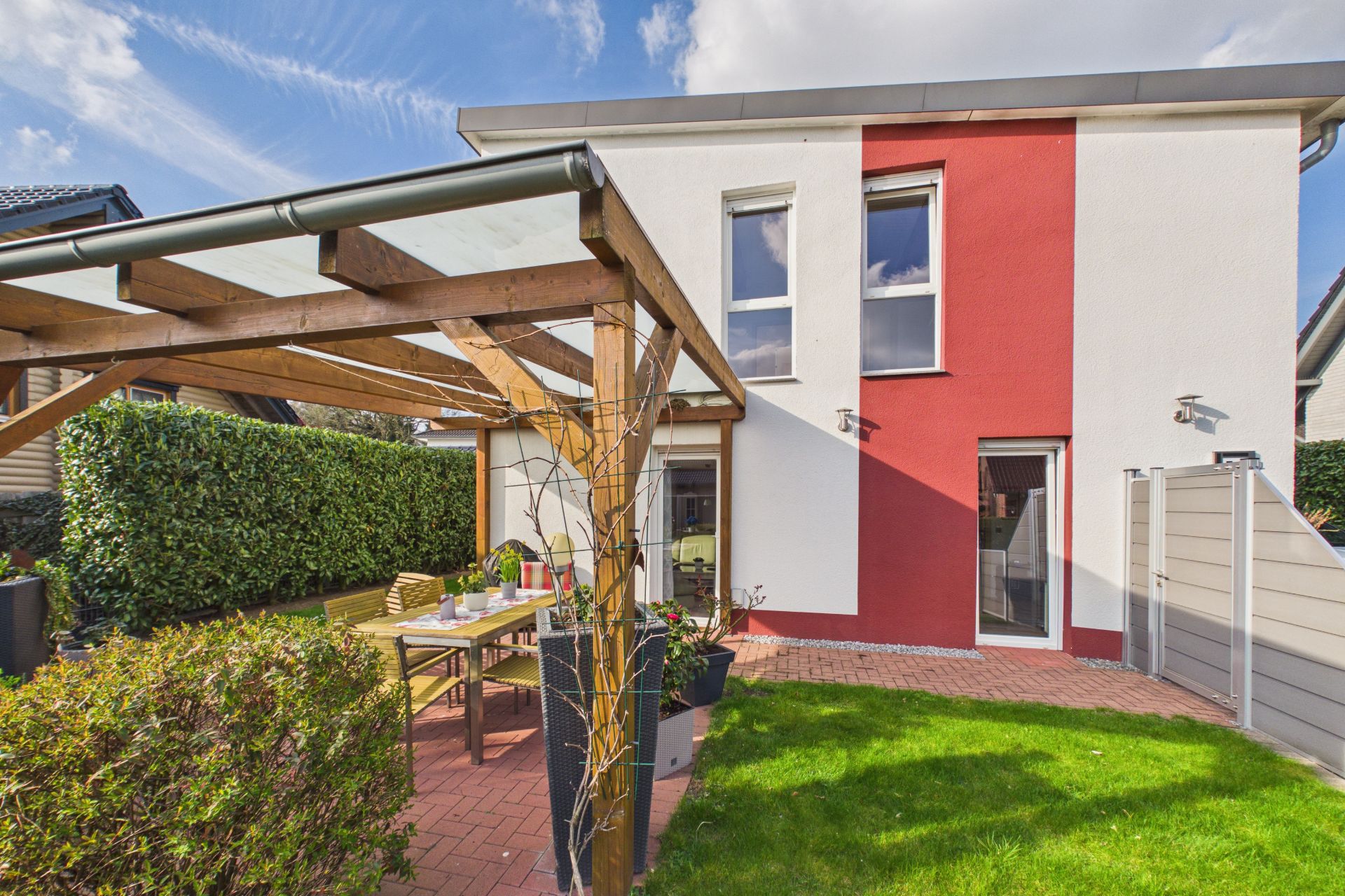 Moderner Garten mit Holzbaldachin, Tisch und Stühlen vor einem zweistöckigen Haus mit roten und weißen Wänden.