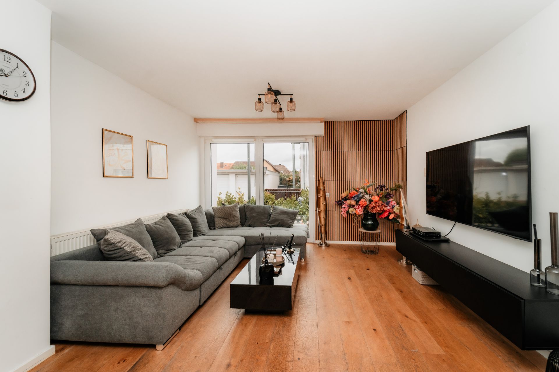 Modernes Wohnzimmer mit grauem Ecksofa, Couchtisch, großem Fernseher und Blumenstrauß.