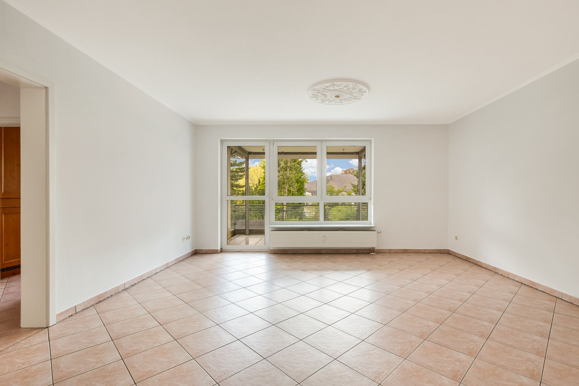 Helles, leeres Wohnzimmer mit Fliesenboden und großem Fenster zur Terrasse.
