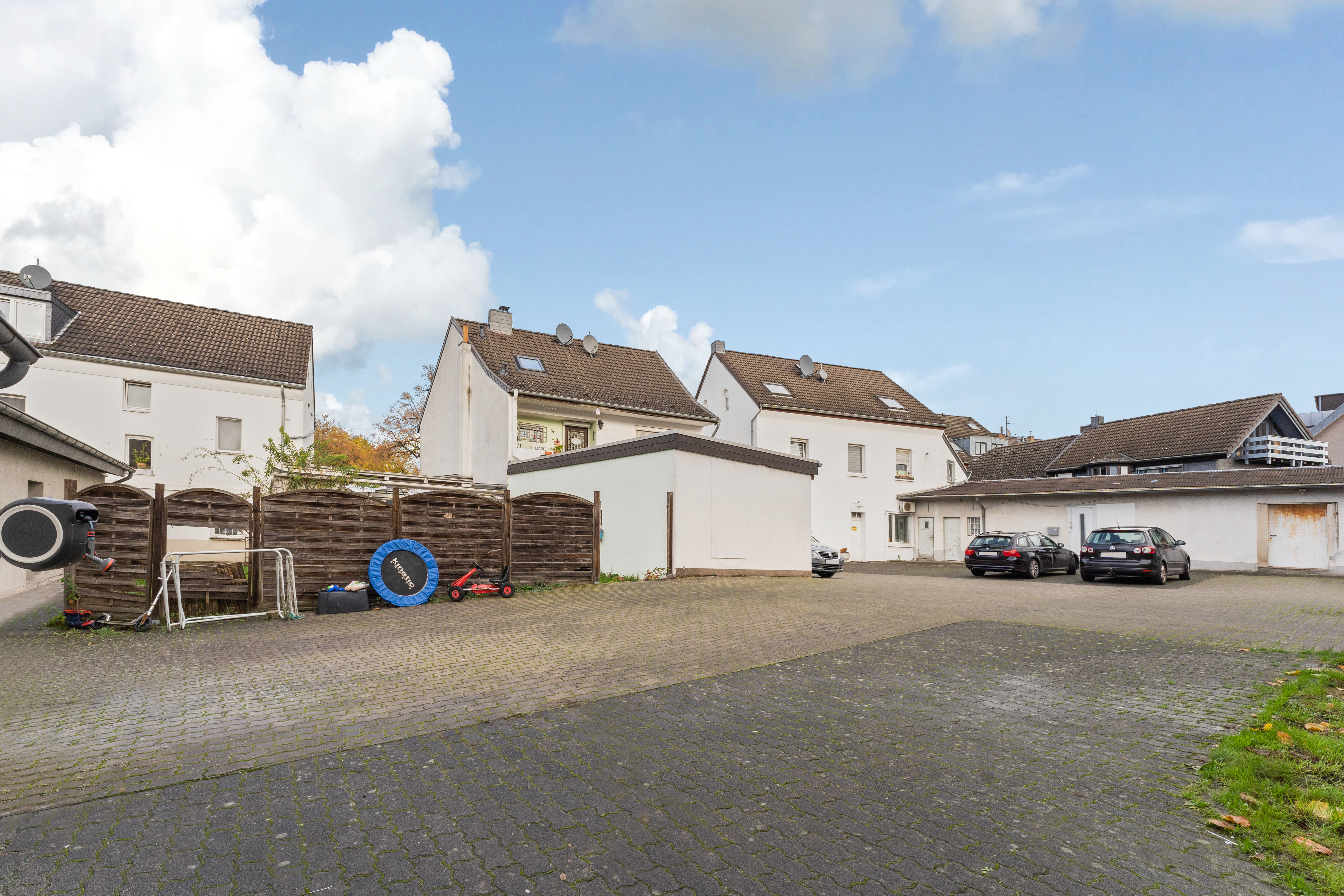 Gepflasterter Hof mit geparkten Autos, Spielgeräten und weißen Wohnhäusern im Hintergrund.