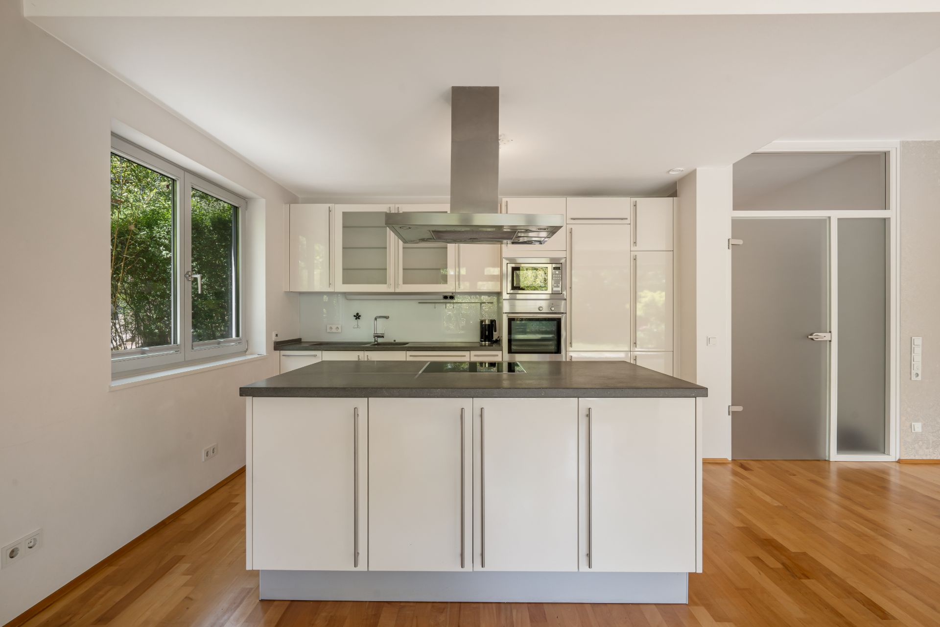 Moderne Küche mit Kochinsel, weißen Schränken und Holzboden, großes Fenster links.