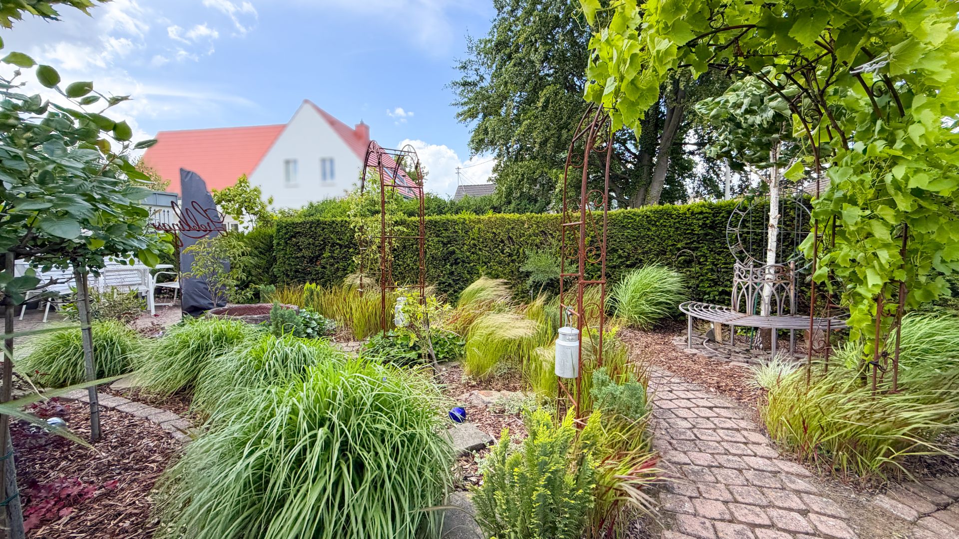Garten mit Pflanzen, Büschen, Sitzbank, Weg aus Pflastersteinen und Rankgittern.