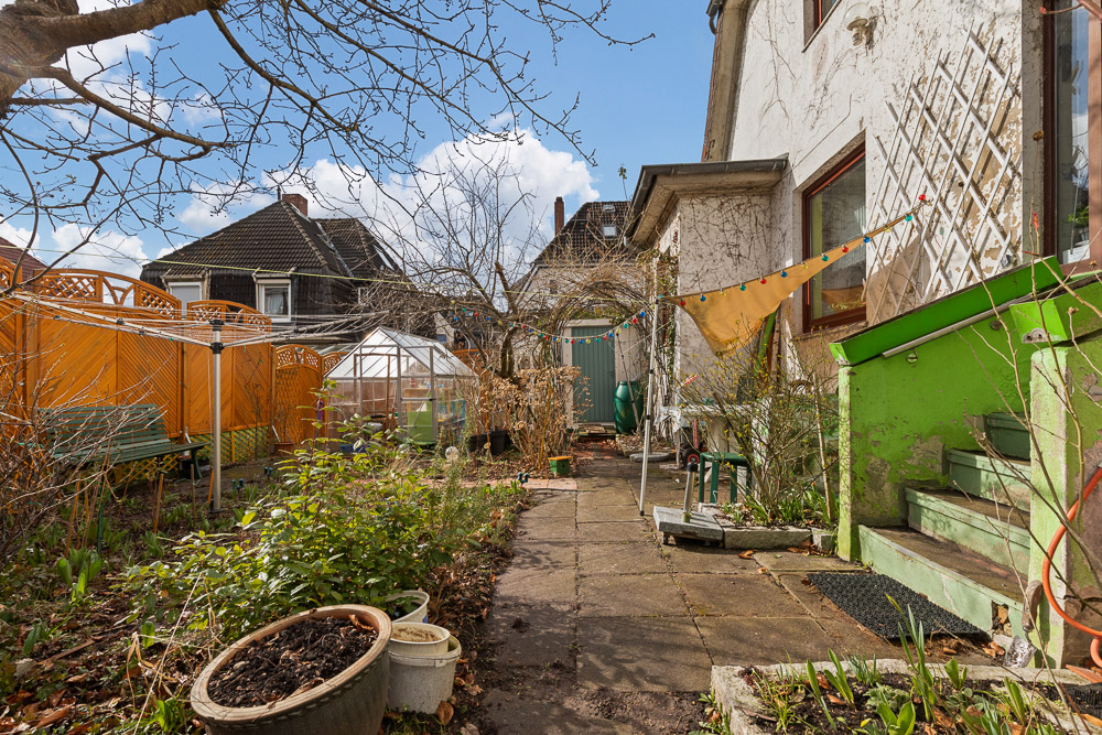 Garten mit Pflanzen, Hochbeet, Gewächshaus, Sitzbank und grüner Treppe neben einem Wohnhaus.